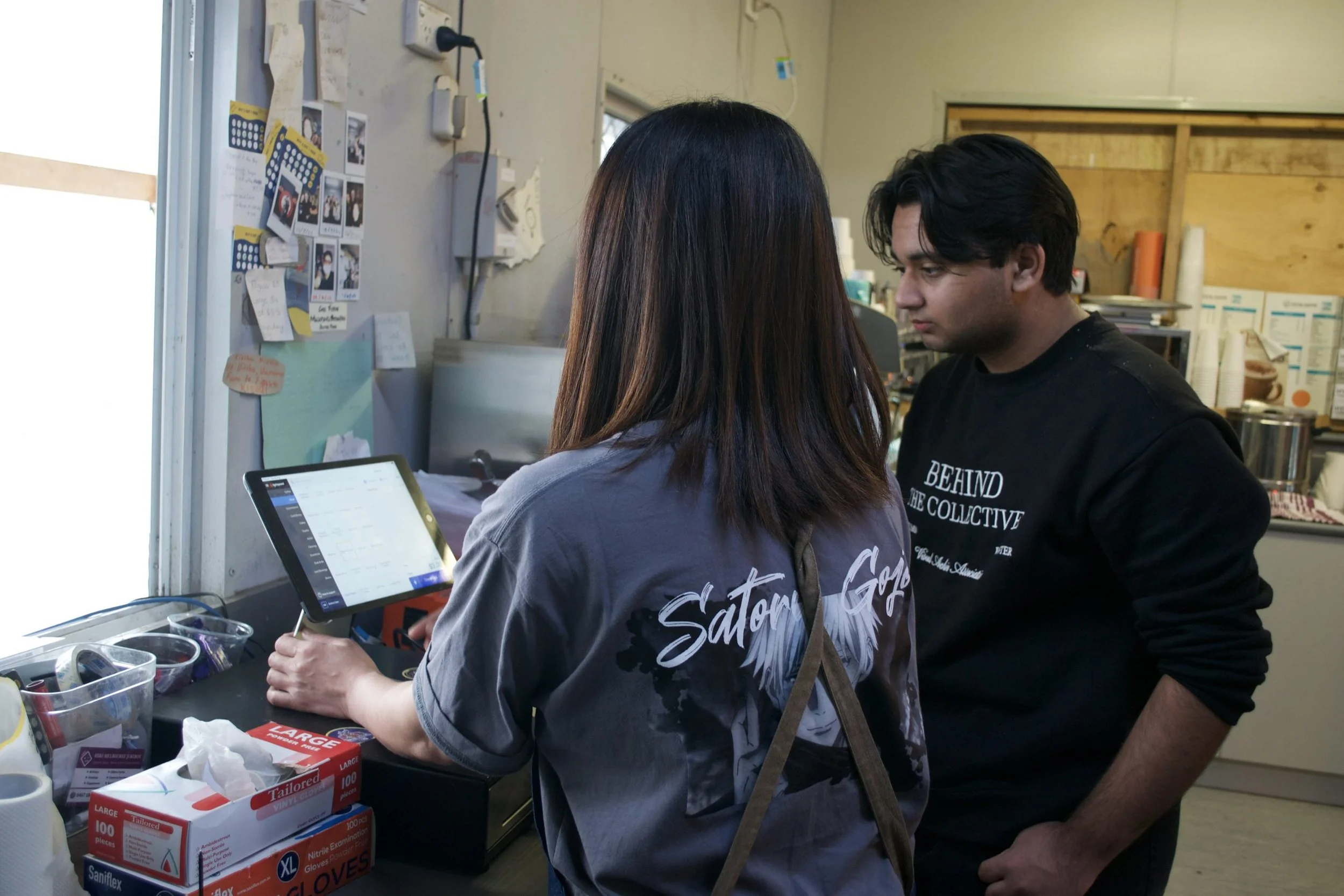 Two people, a woman and a man, standing at a counter in a small kitchen or food stand. The woman is using a tablet, and the man is standing close, looking at the screen. The woman is wearing a gray shirt with a graphic and the words "Sator G..." on the back. The man is wearing a black sweatshirt with white text that says "BEHIND THE COLLECTIVE." The counter has some supplies, including a box of large gloves, and the background shows a cluttered kitchen with various items.