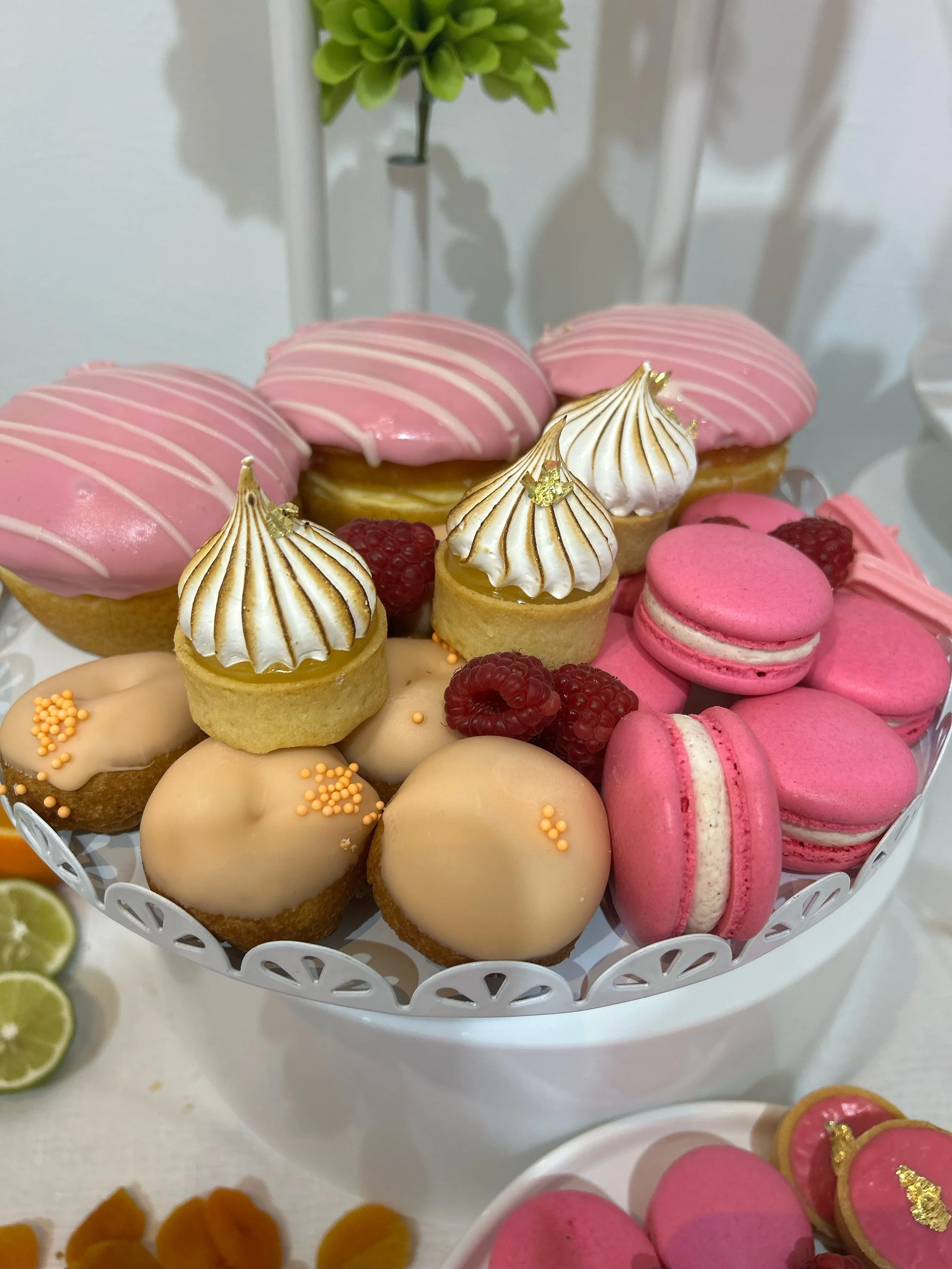 A white decorative cake stand holding assorted pink and white desserts, including macarons, small tarts with meringue peaks, and mini eclairs, topped with gold leaf and sprinkles, garnished with raspberries.