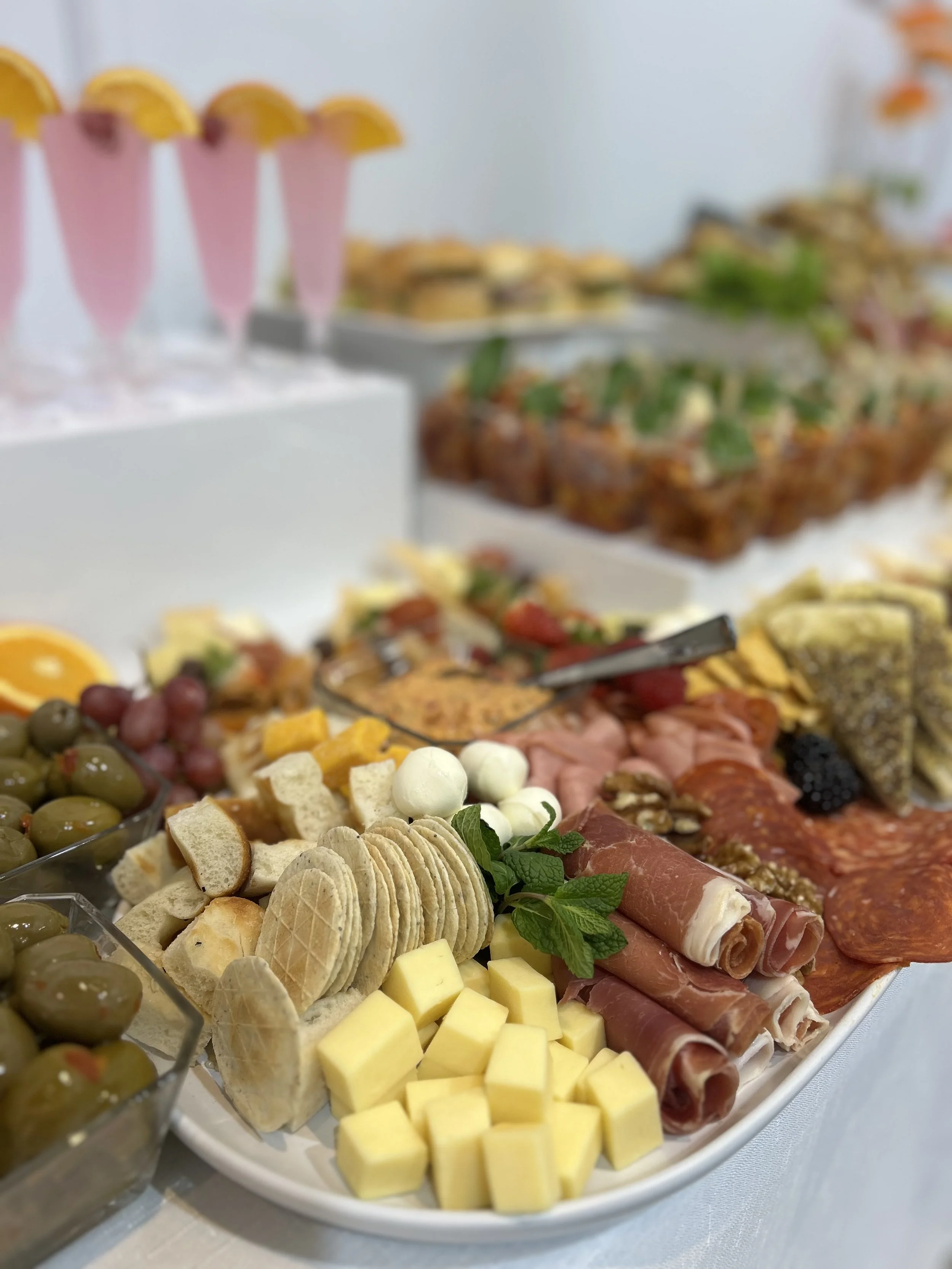Charcuterie platter with cheese cubes, sliced meats, crackers, grapes, olives, and garnishes, with Italian soda in pink cups in the background.