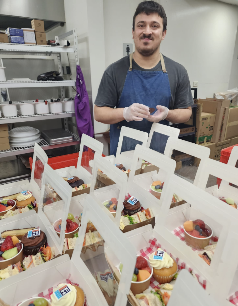 A man wearing gloves and an apron stands behind a table filled with assorted desserts, including cupcakes and fruit cups, in a storage or kitchen area.