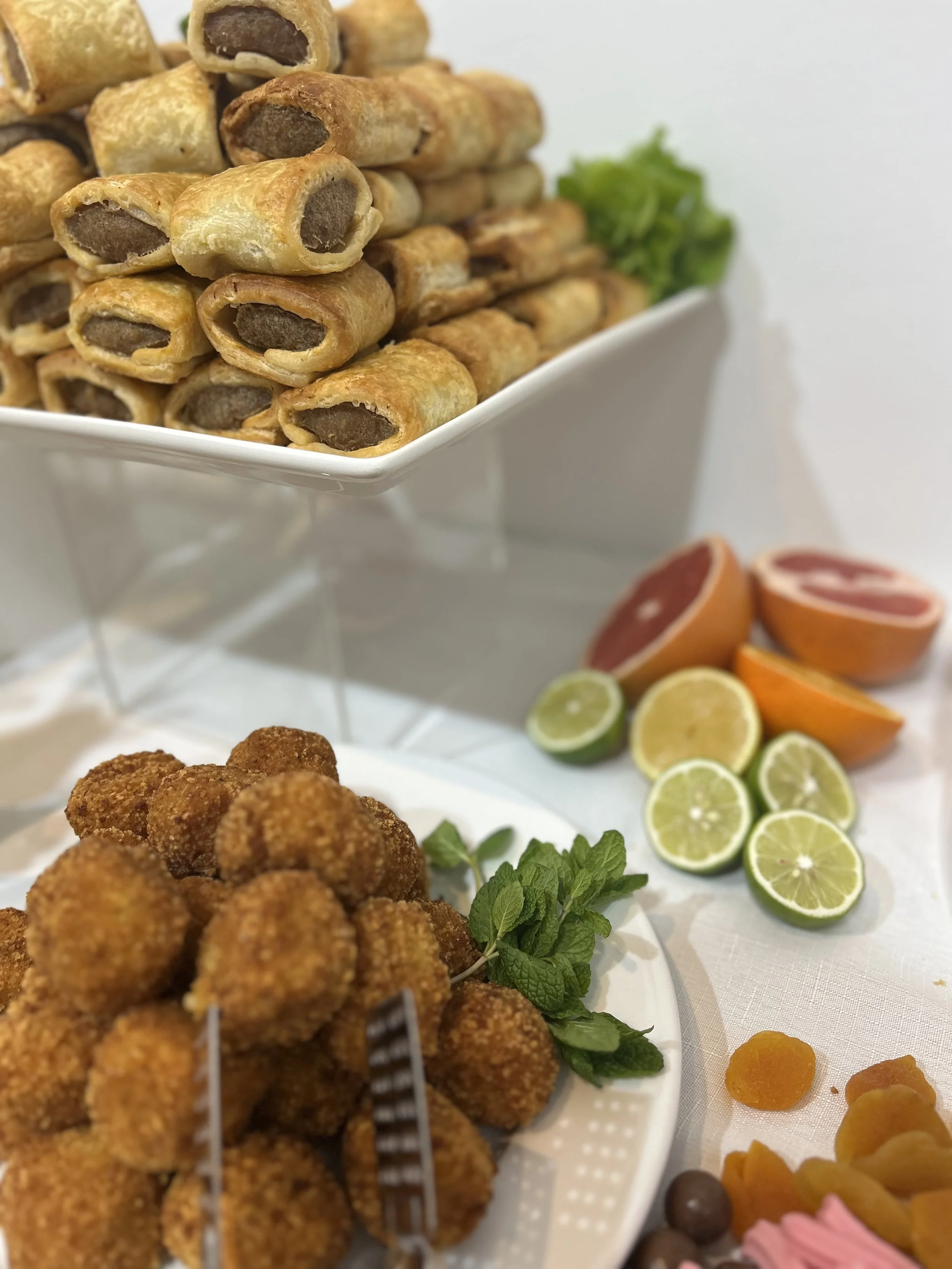 Plate of breaded fried appetizers, sliced citrus fruits, and fresh herbs with a stack of egg roll appetizers in the background.