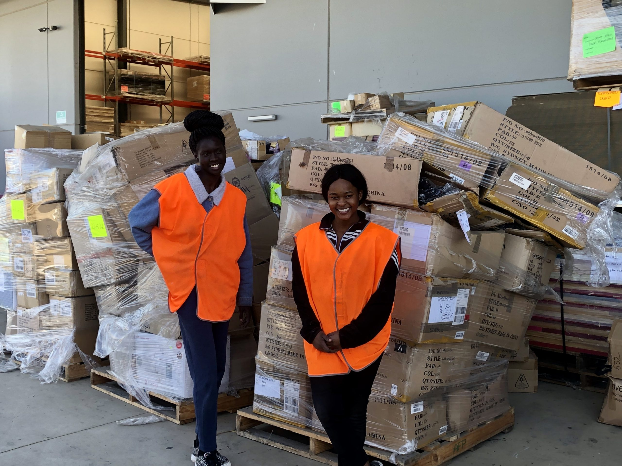 Two women wearing orange safety vests standing in front of stacked and wrapped boxes near a warehouse wall.