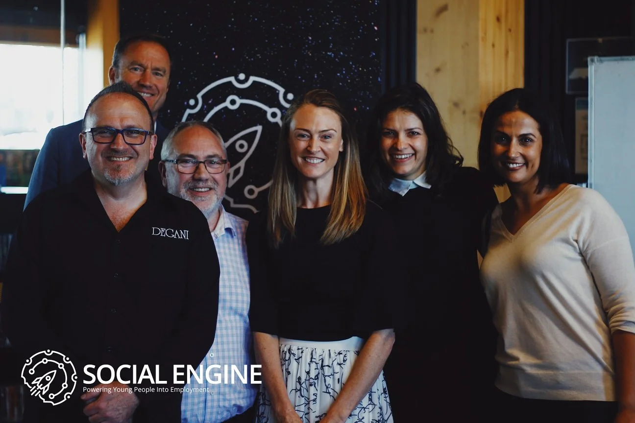 Group of six diverse professionals smiling indoors at a social or corporate event, with a logo and text "SOCIAL ENGINE - Powering Young People Into Employment" in the lower left corner.