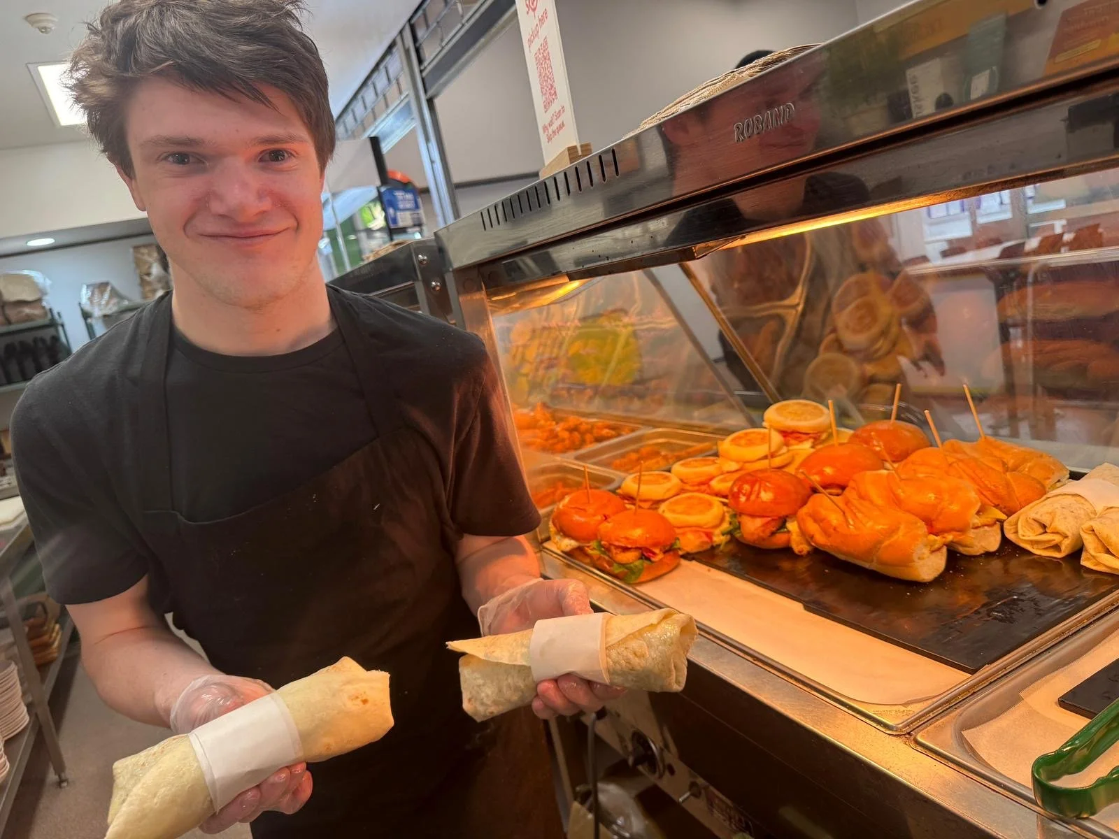 A young man with short dark hair, wearing a black T-shirt and gloves, is holding a wrapped sandwich or burrito and standing next to a counter with cooked sandwiches or sliders in a deli or restaurant. The man is smiling, and behind him are trays of s