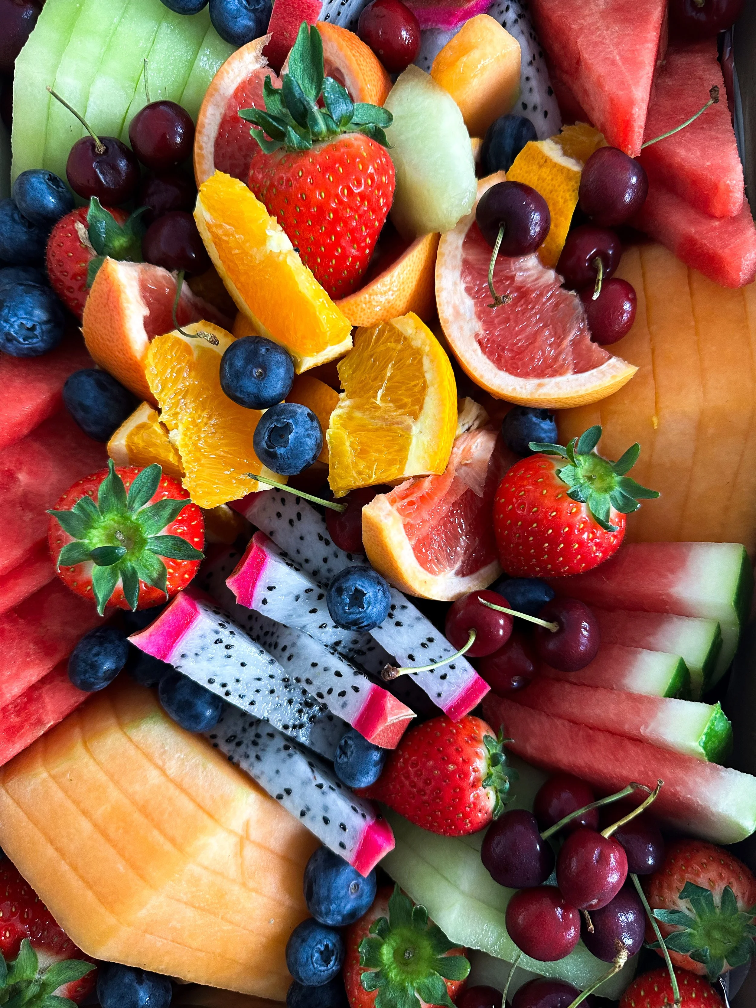 Assorted sliced fruits including strawberries, blueberries, watermelon, honeydew, cantaloupe, grapefruit, orange, dragon fruit, and cherries.