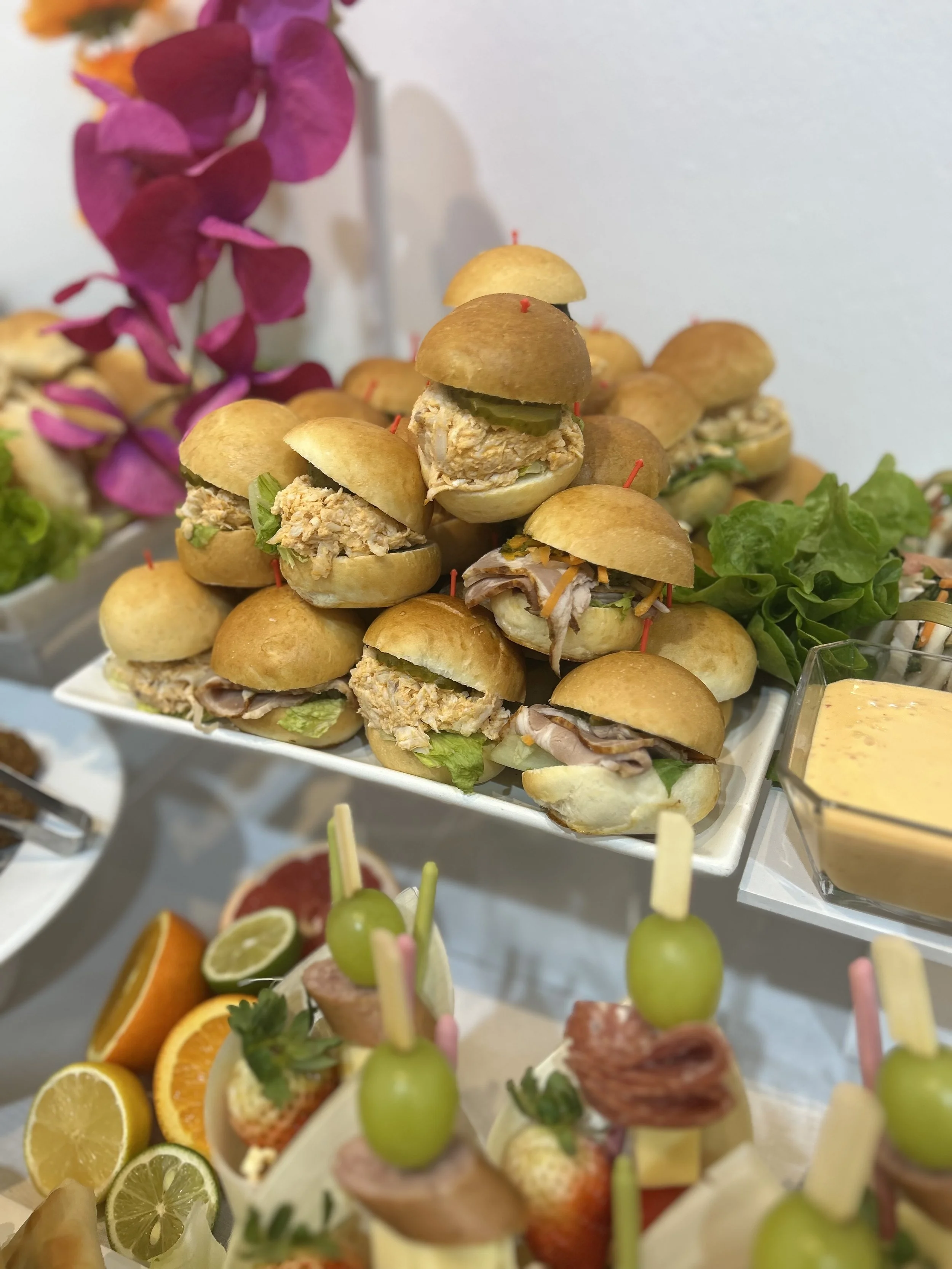Mini sandwiches with chicken, tuna, and ham, garnished with lettuce, pickles, and peppers, arranged on a white platter, with colorful fruit skewers and a side of dipping sauce, and decorated with pink flowers in the background.