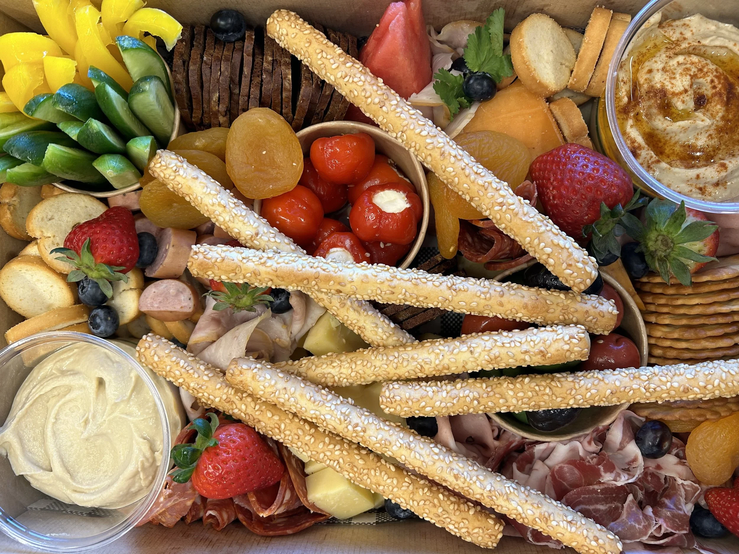 A platter with assorted fresh fruits, cheese, crackers, and breadsticks, including strawberries, blueberries, grapes, cherry tomatoes, apricots, and melon, garnished with herbs and served with dips and spreads.