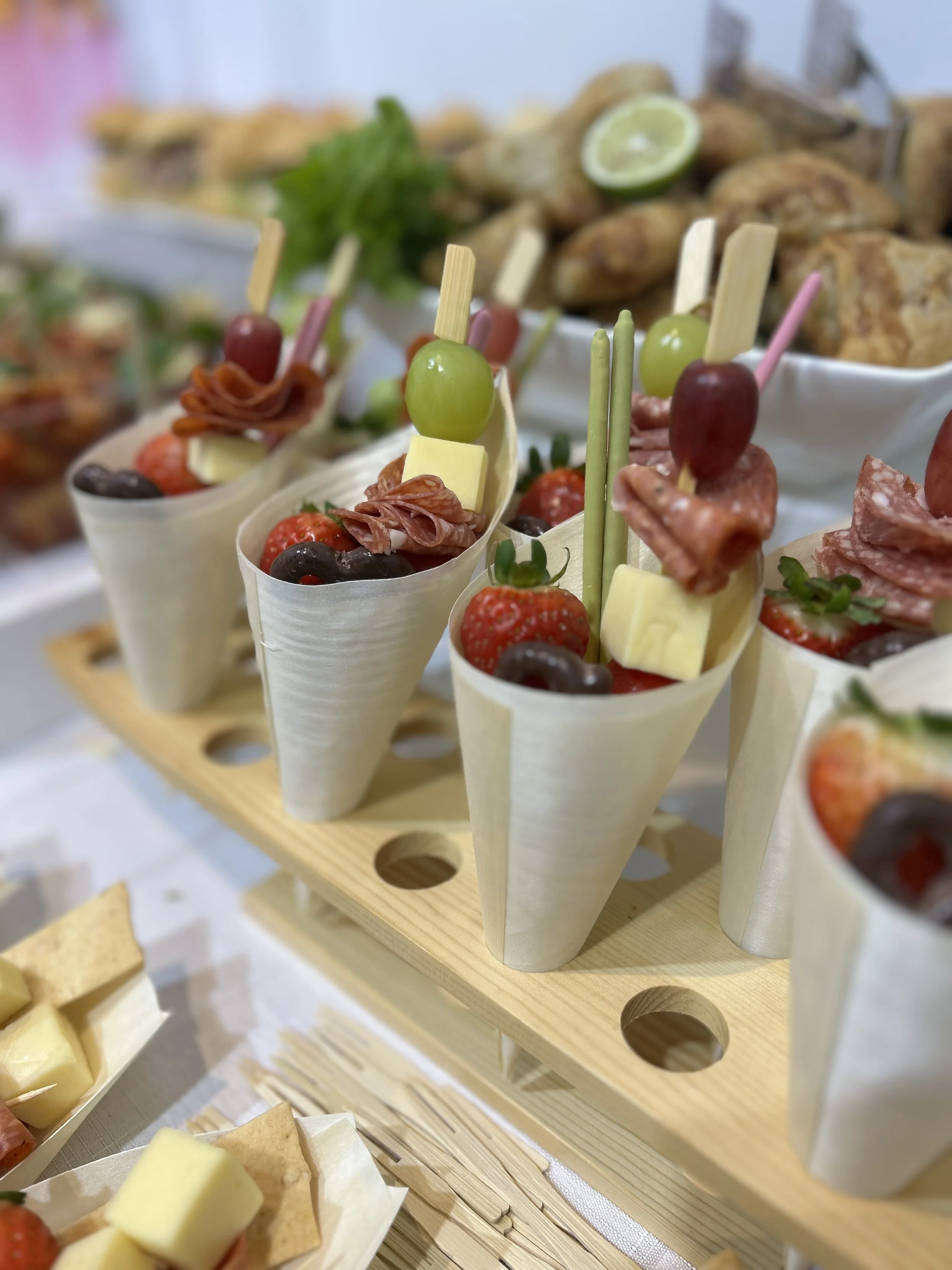 Assorted fruit and cheese appetizers in cone-shaped containers on a wooden platter, topped with grapes, strawberries, cherry tomatoes, and slices of cheese.