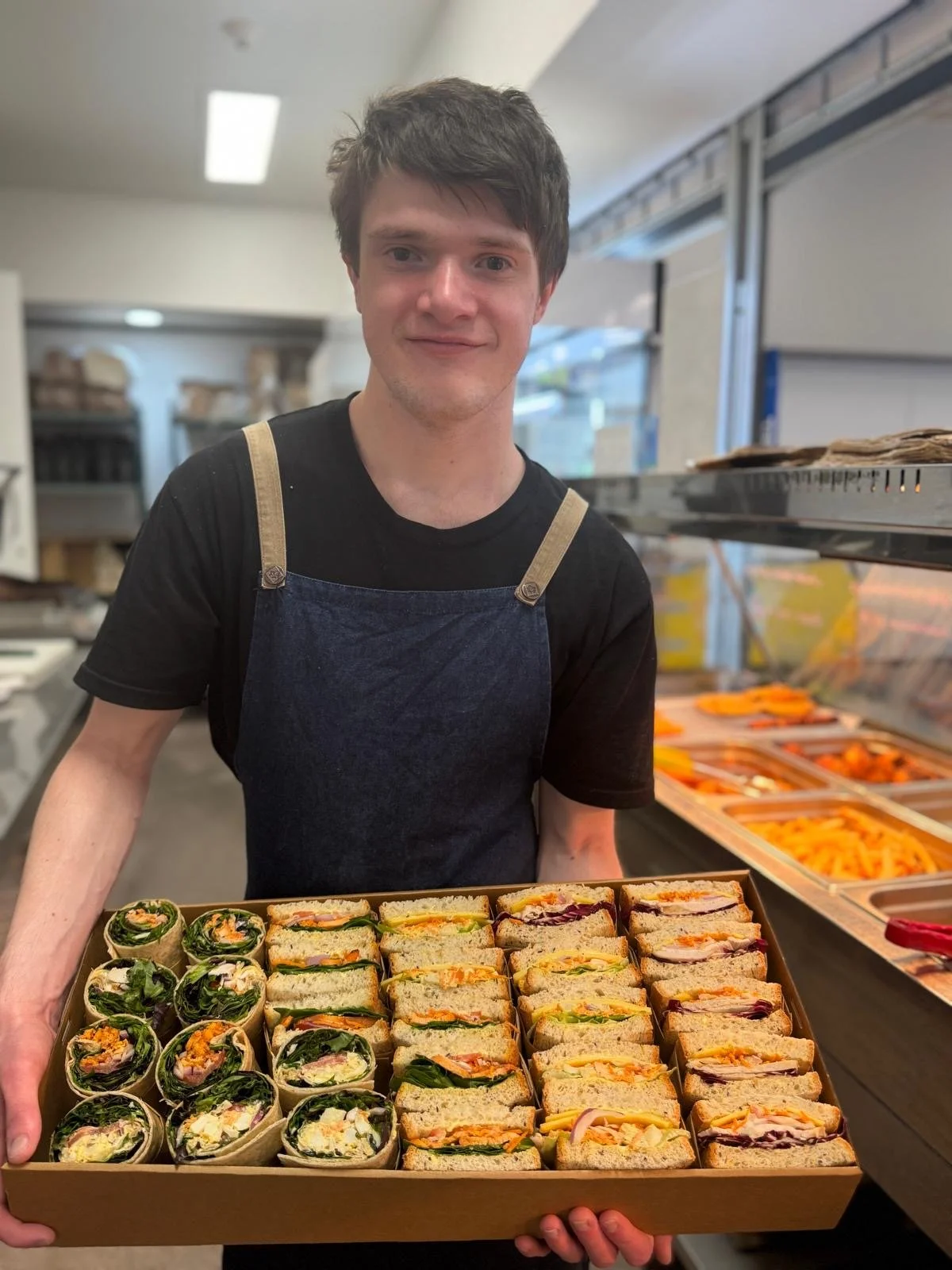 A young man wearing a black t-shirt and denim apron holding a tray of assorted sandwiches inside a store or deli.
