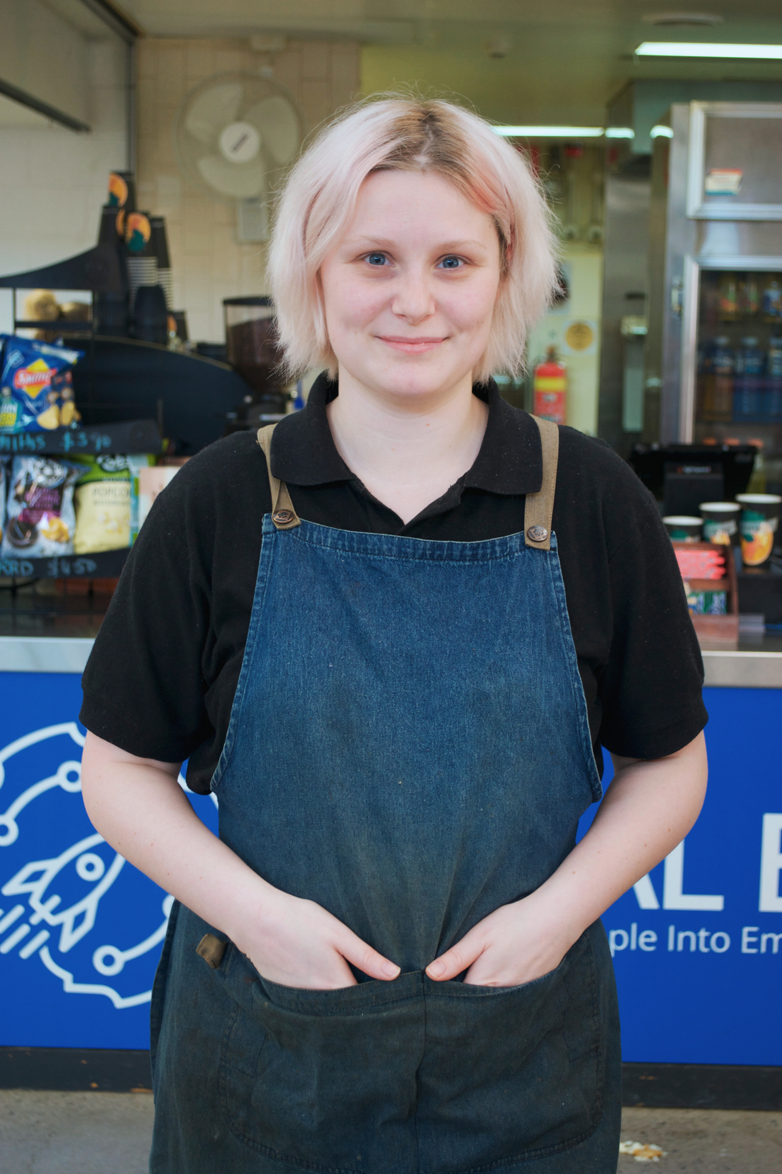 A young woman with pinkish blonde hair, wearing a black polo shirt and a denim apron, standing in front of a counter at a cafe or snack bar, smiling at the camera.
