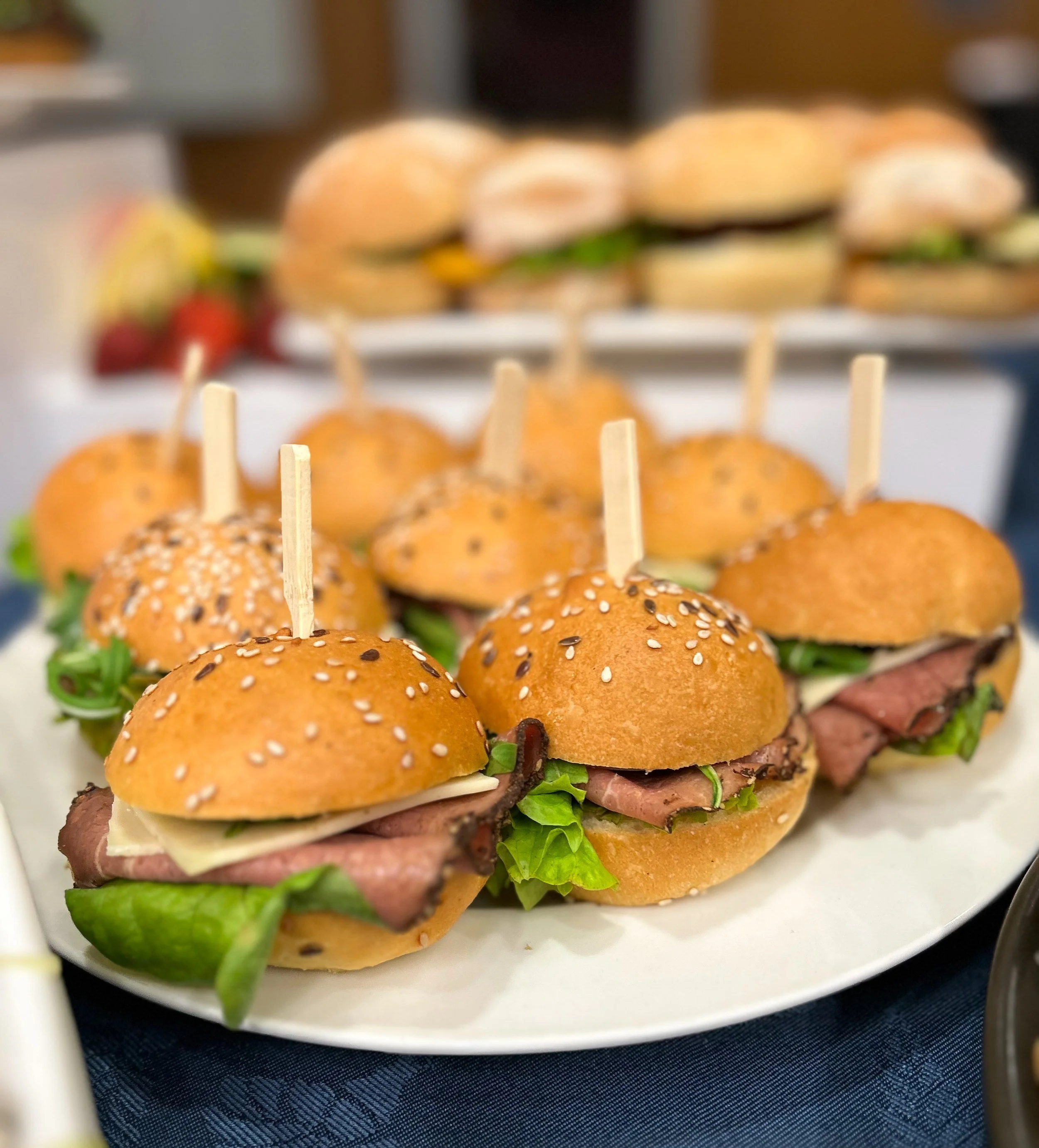 Platters of mini sandwiches with roast beef, lettuce, and cheese on sesame seed buns, held together with wooden toothpicks.