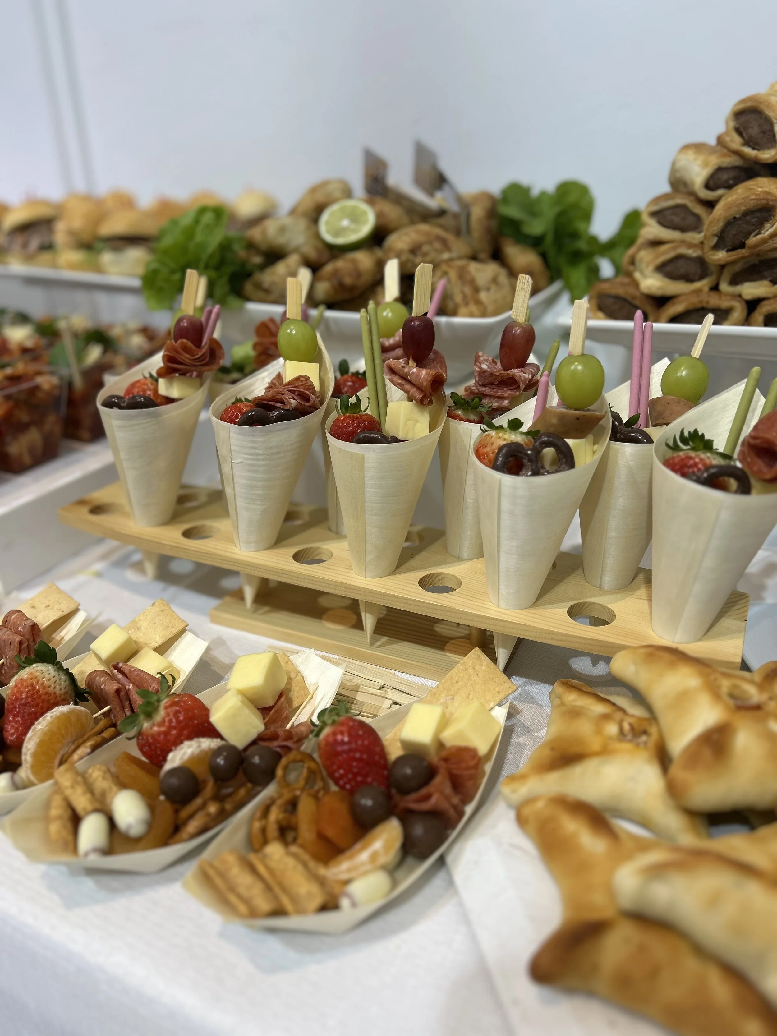 A buffet table with assorted food items including fruit cups with strawberries, grapes, and cheese, pastries, and small sandwiches or appetizers, with a background of more finger foods and pastries.
