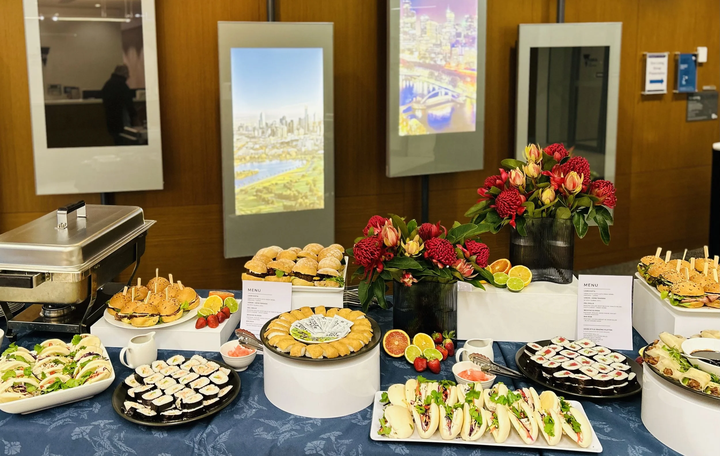 A buffet table with assorted sandwiches, sushi, and pastries, decorated with red and yellow flowers, alongside fresh citrus fruits, in front of a display of framed artworks on a wooden wall.