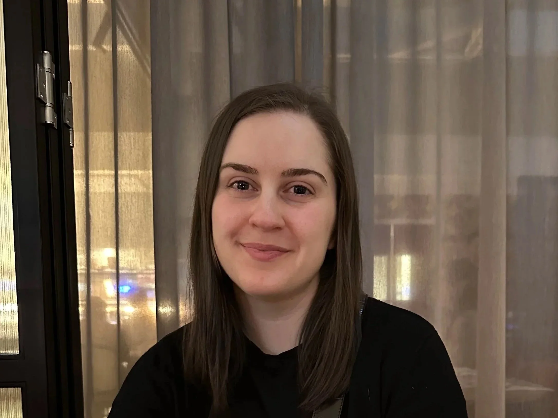 A young woman with long brown hair, smiling, in front of sheer curtains and a glass door.