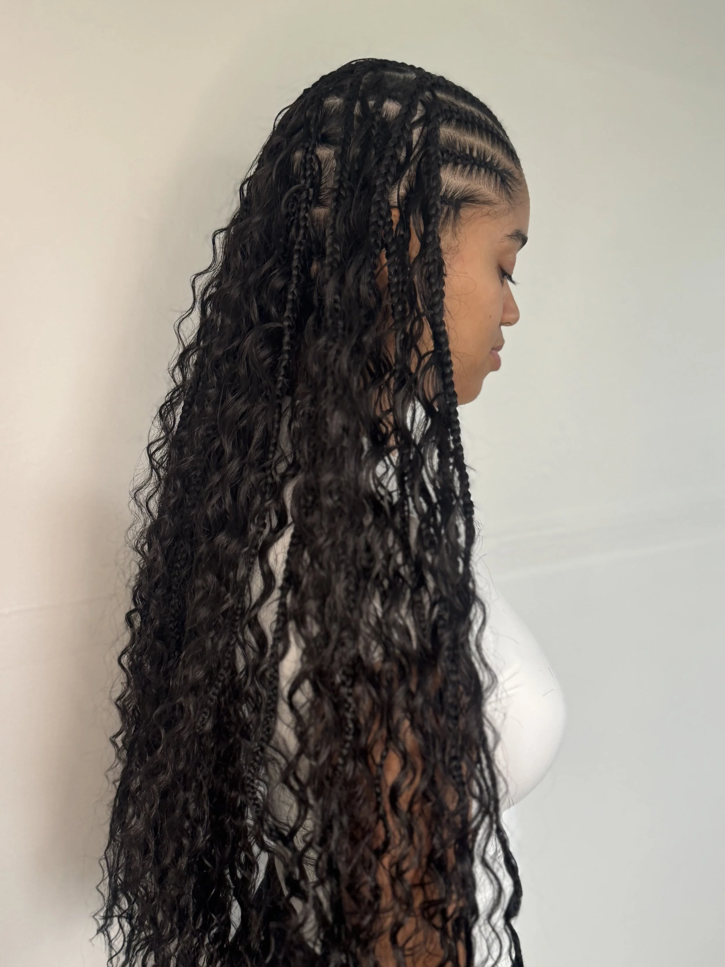 A woman with long, curly, braided hair standing sideways against a plain white wall.
