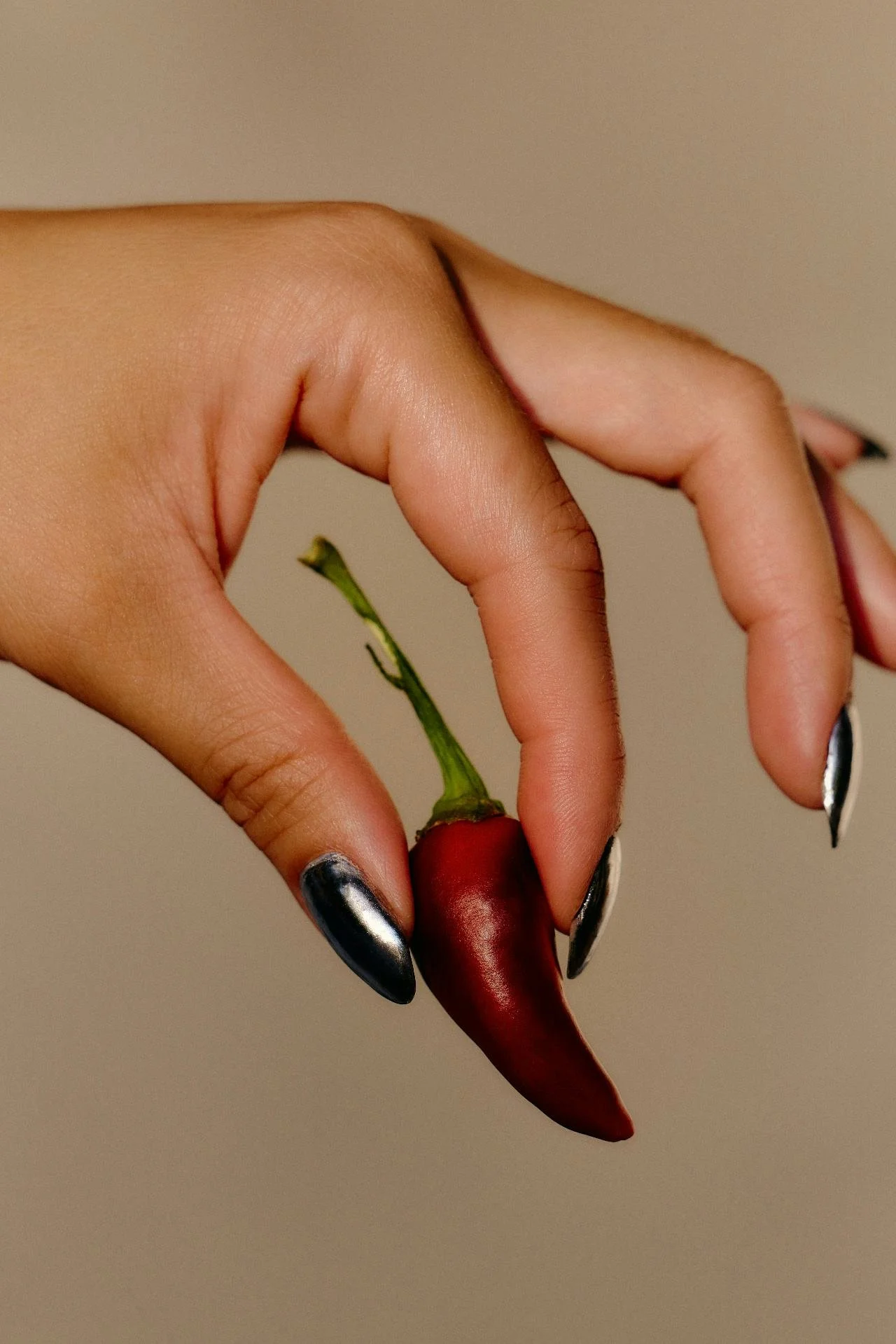 A hand with metallic black painted nails holding a small red chili pepper with a green stem.