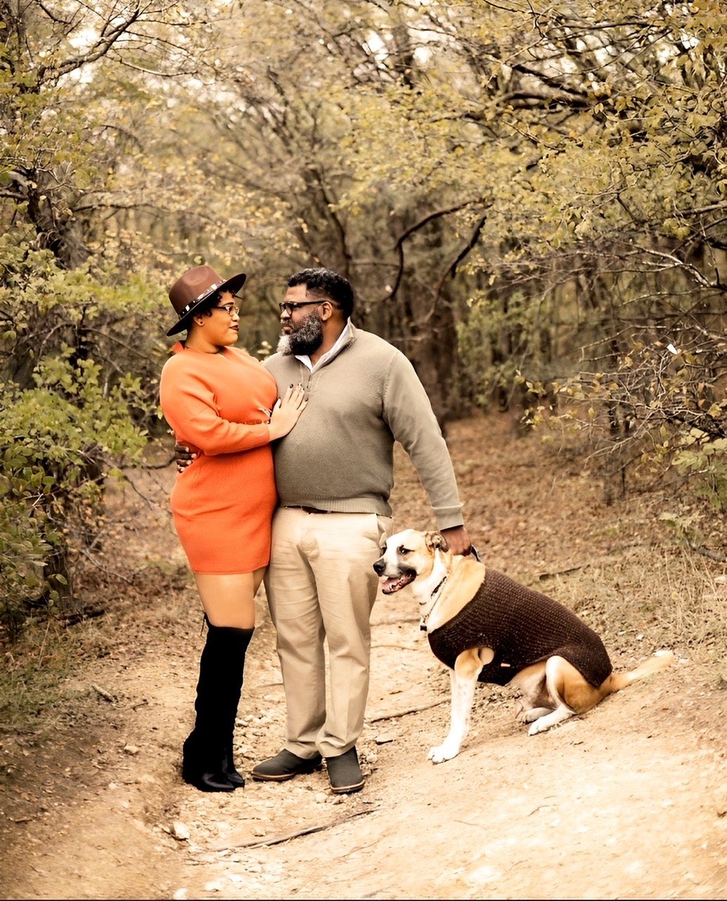 A couple standing on a forest trail, looking into each other's eyes, with a dog wearing a sweater sitting nearby. The woman wears an orange dress, black knee-high boots, glasses, and a brown hat. The man wears a beige sweater, beige pants, and glasses. The scene appears to be during fall with trees displaying yellow and brown leaves.