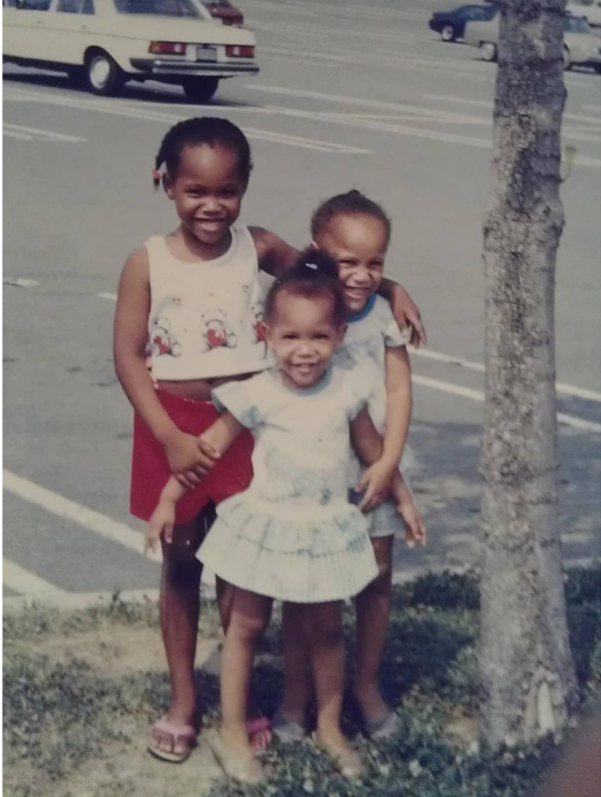 Three young girls smiling and standing together outdoors near a tree, with cars parked and a parking lot in the background.