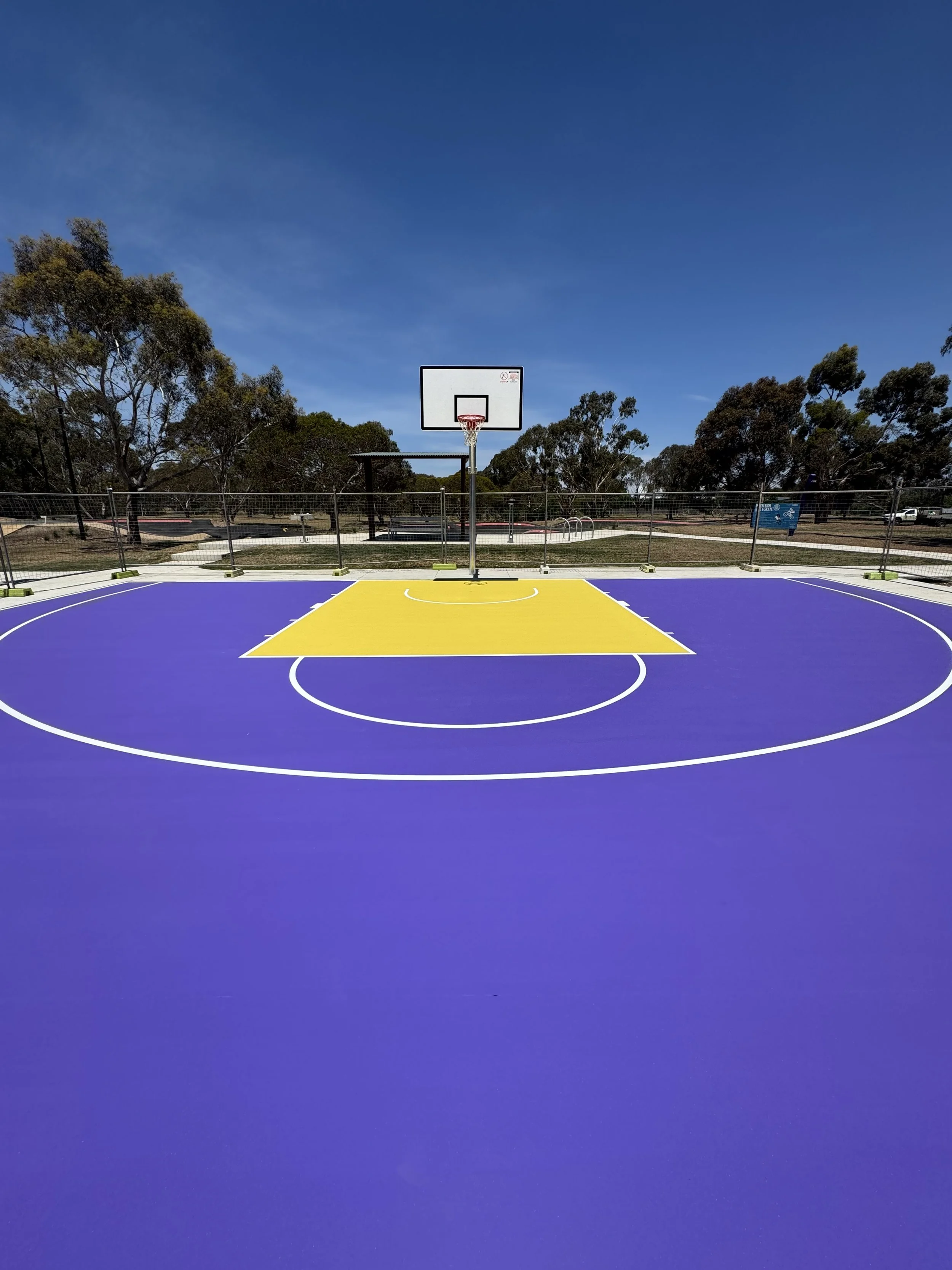 An outdoor basketball court with a purple surface and yellow key area, surrounded by a fence, against a background of trees and a blue sky.