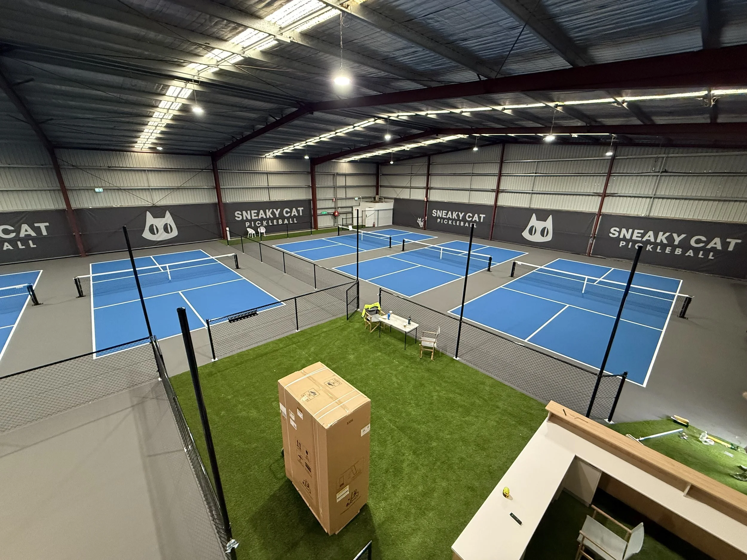 Indoor pickleball courts with blue surfaces, surrounded by black netting and walls with 'Sneaky Cat Pickleball' branding, overhead lighting, and some chairs and a cardboard box nearby.