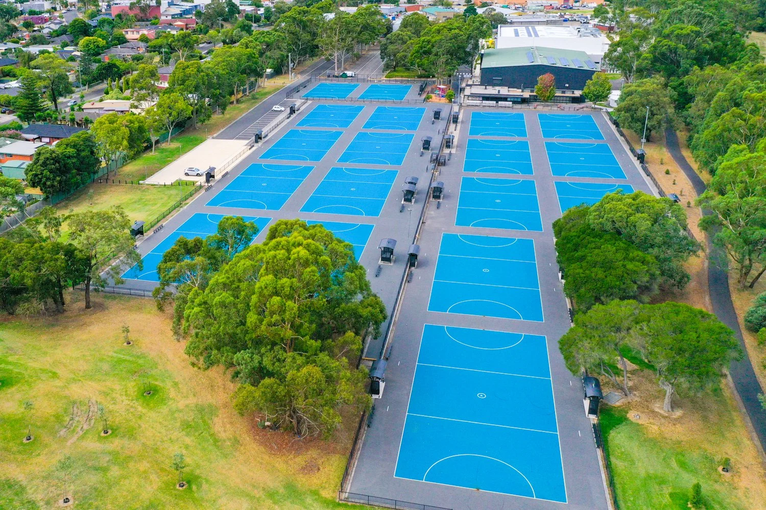 Aerial view of a sports complex featuring multiple blue basketball courts with white lines, surrounded by trees and greenery, with residential and commercial buildings in the background.