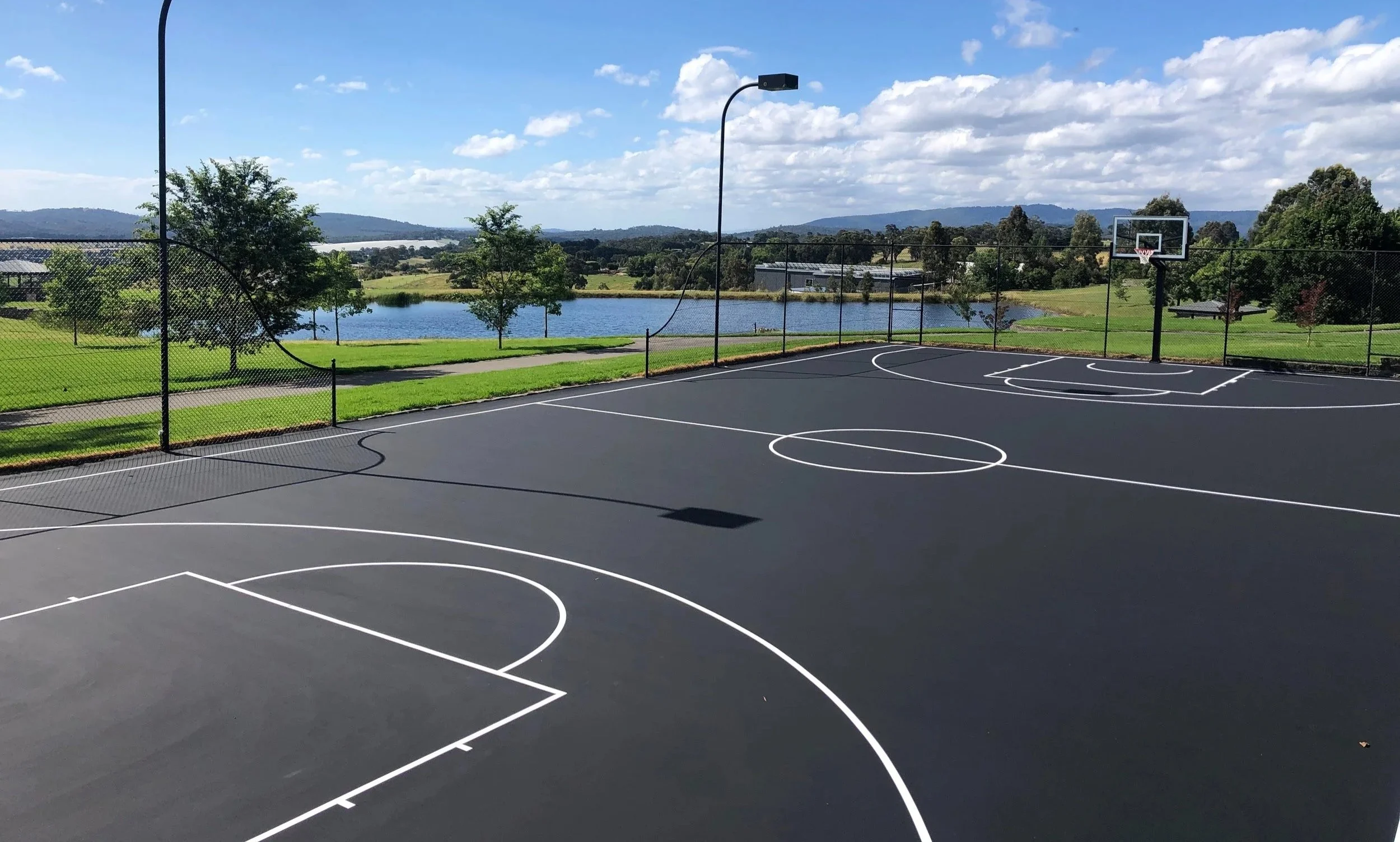 Empty outdoor basketball court with a black surface, white lines, and two hoops, surrounded by a fence, with a lake, trees, and hills in the background under a partly cloudy sky.