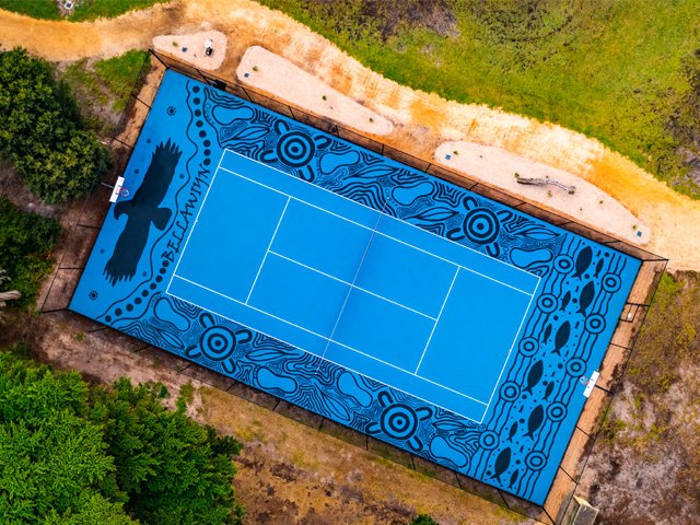 A top-down view of an outdoor tennis court with blue surface, surrounded by trees and dirt paths, featuring Aboriginal-style artwork and a black eagle silhouette, with the word "BLAKAWIN" written on one side.