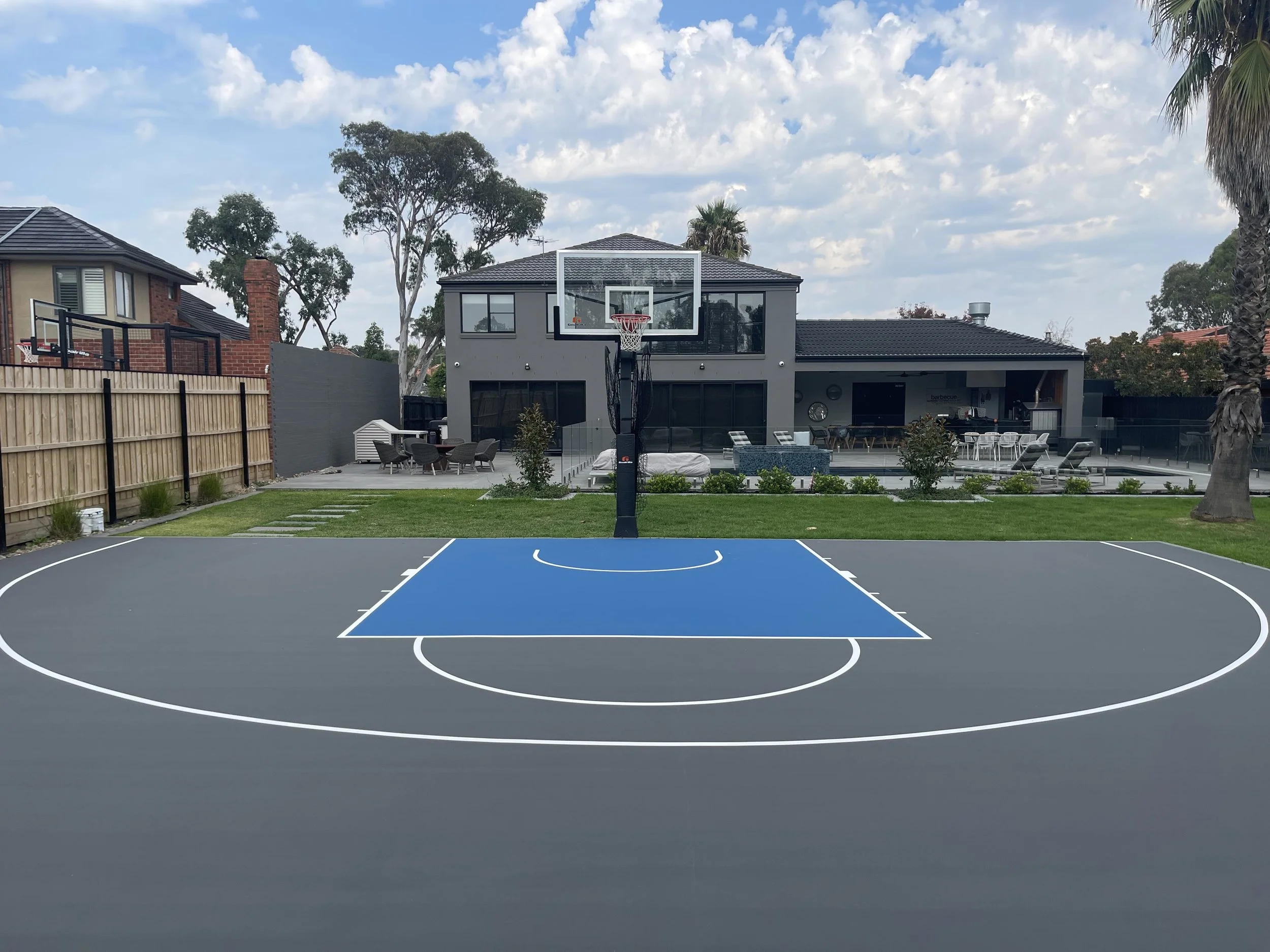 Backyard basketball court with a house in the background, patio furniture, and tall trees.