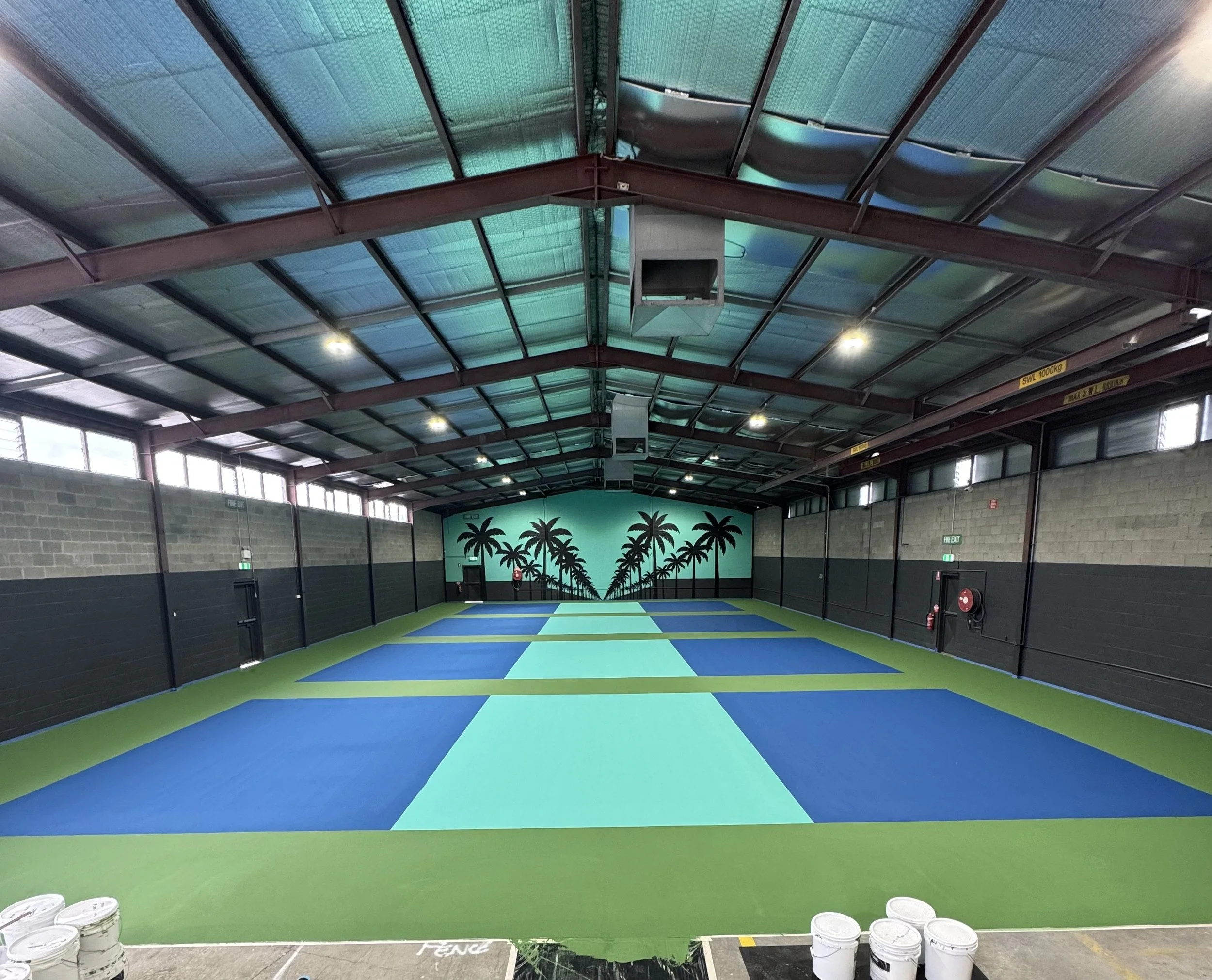 Indoor sports facility with a bright green and blue court, palm tree mural on the back wall, and a high arched ceiling with lighting.