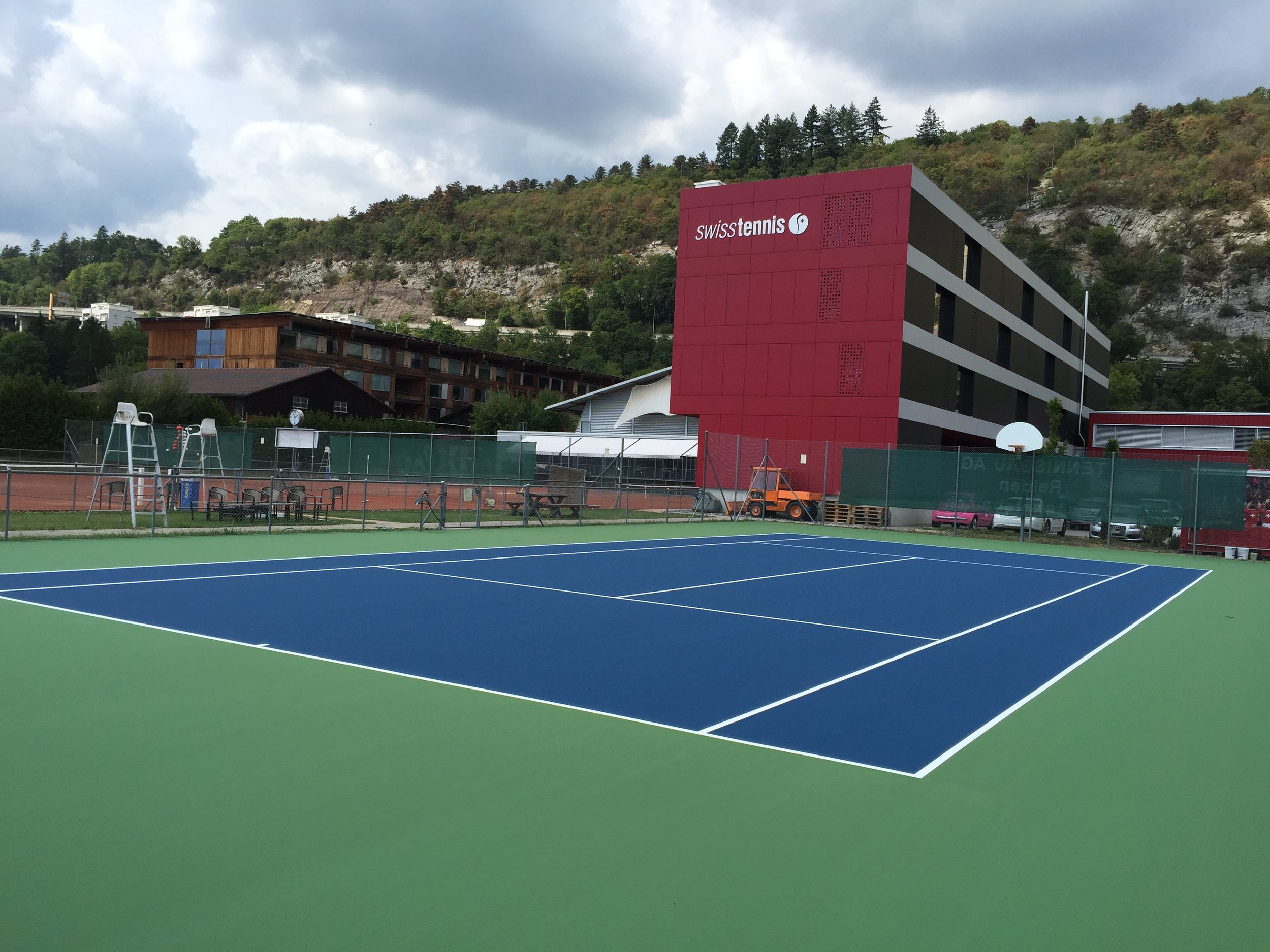 An empty tennis court with a blue playing surface and green outer area, enclosed by a green fence. In the background, there are picnic tables, a playground, and a red building with the sign 'swisstennis'. Hills covered in trees are behind the building, under a cloudy sky.