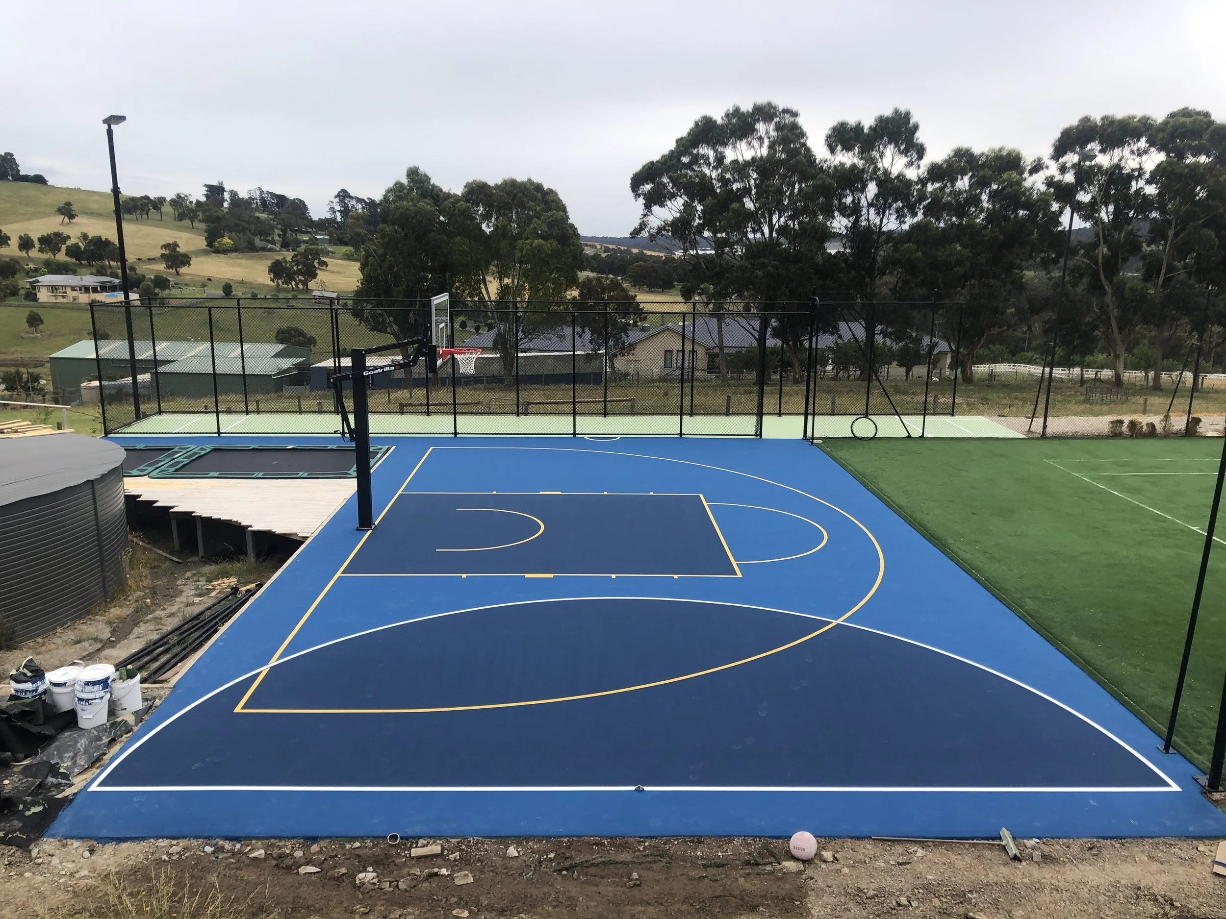 Newly painted outdoor basketball court with blue surface, black safety fence, and surrounded by green grass and trees in the background.
