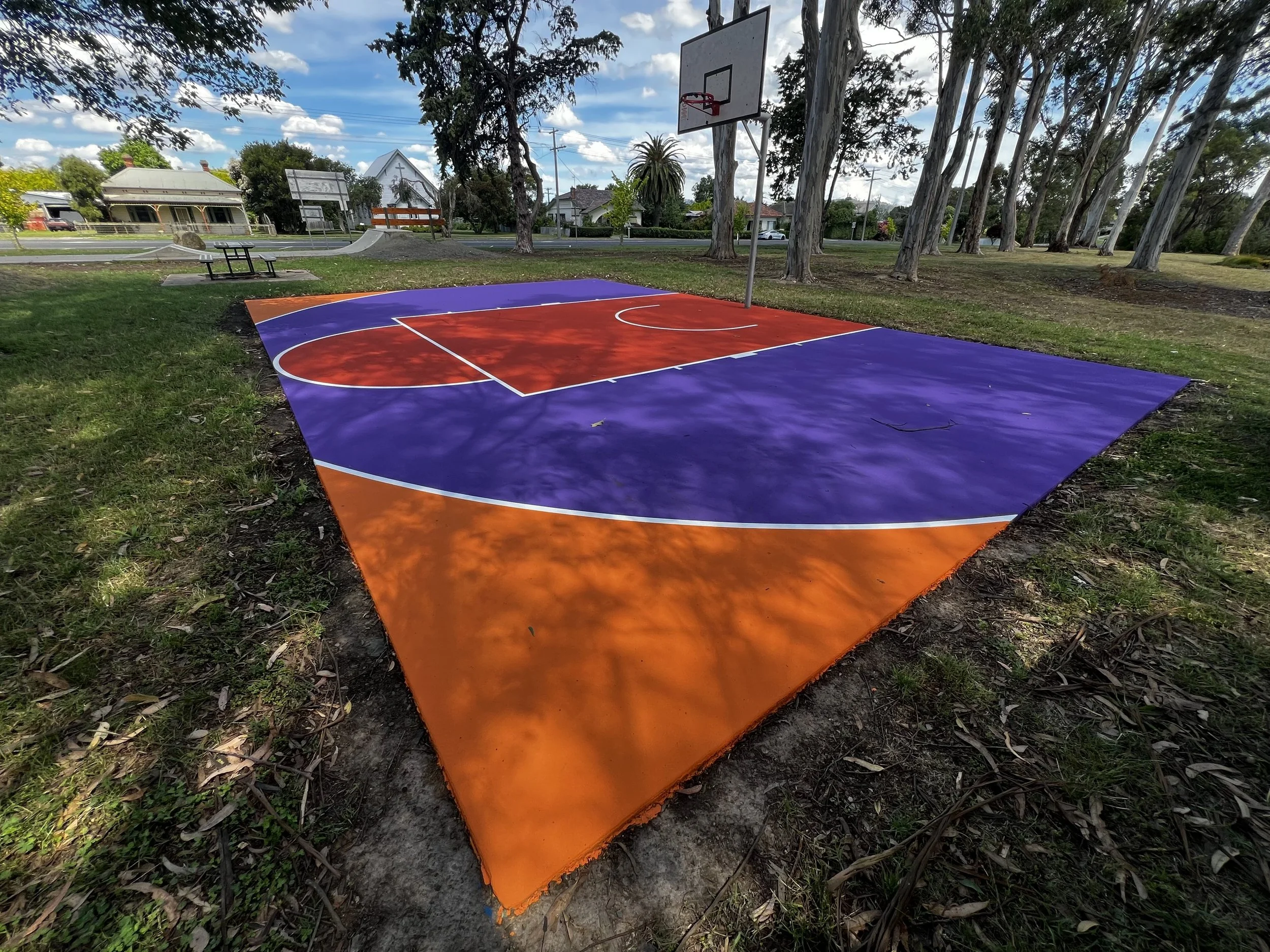 Colorful outdoor basketball court with purple and orange surface, basketball hoop, surrounded by trees and residential houses under a partly cloudy sky.