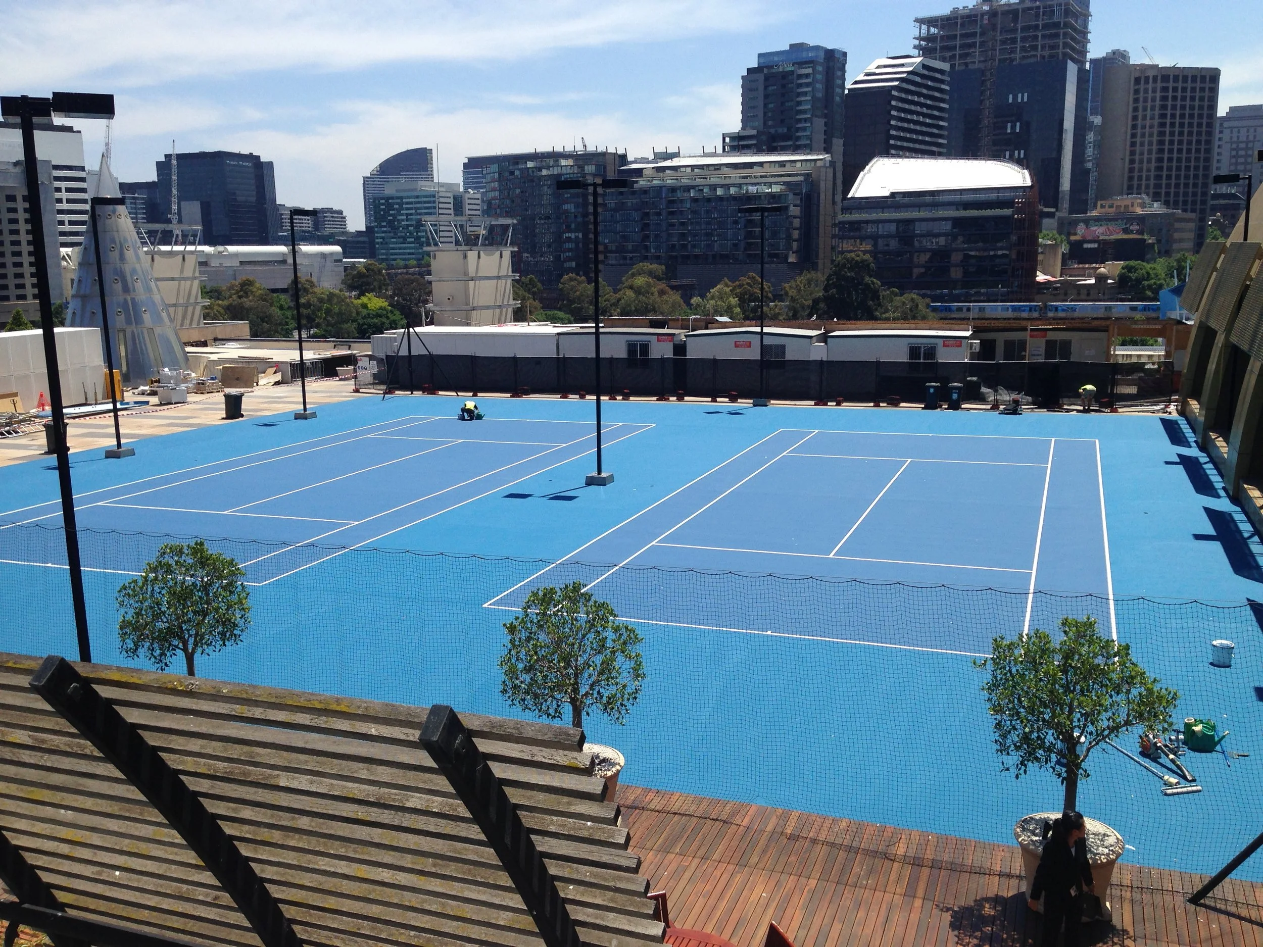 An outdoor rooftop tennis court with a bright blue surface, yellow and white lines, surrounded by small green trees and urban city buildings in the background.