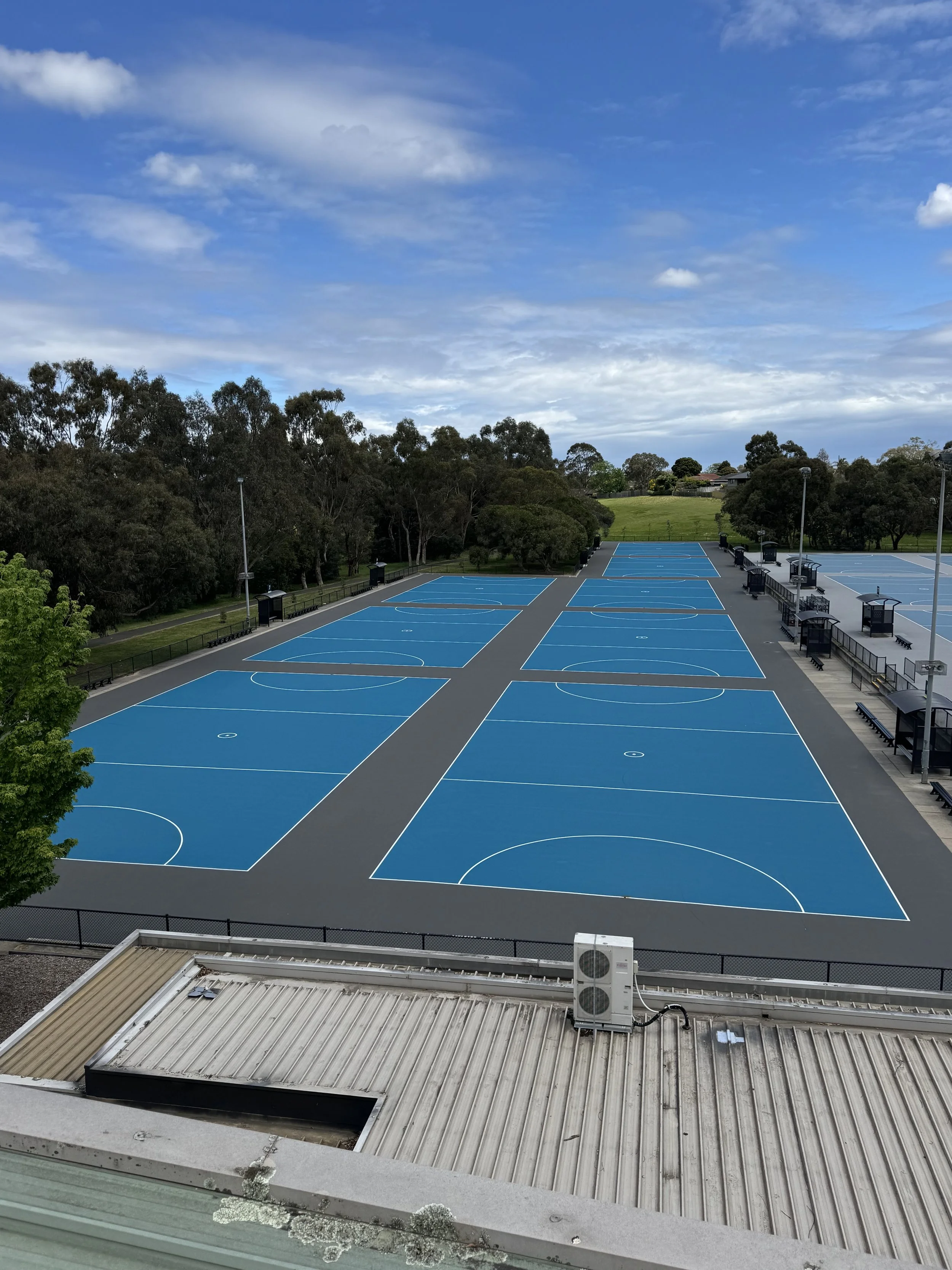Empty blue outdoor basketball courts with white lines, surrounded by green trees and a partly cloudy sky.