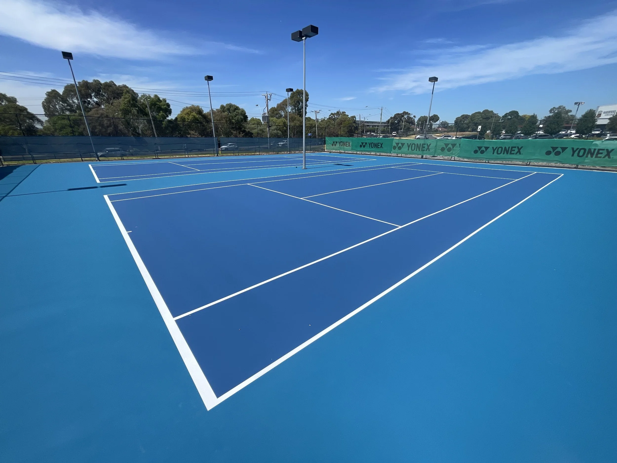 Empty blue tennis court with white lines and net, surrounded by green wind screens with Yonex logo, under a clear blue sky.