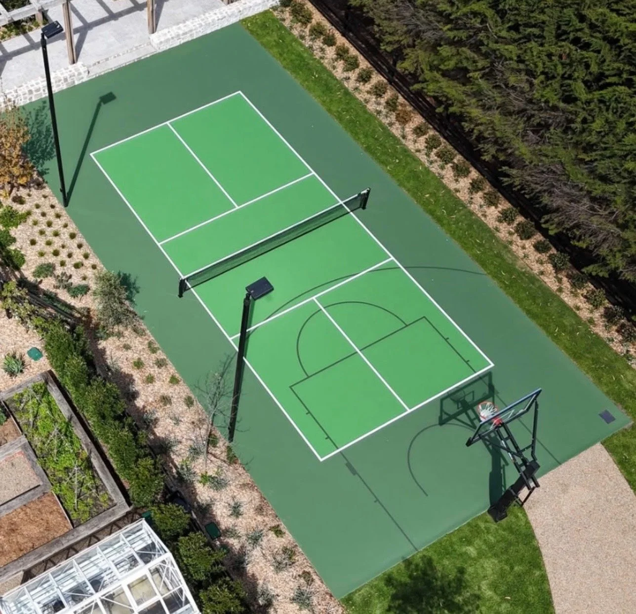 An aerial view of a green outdoor basketball and tennis court, with two basketball hoops at each end and a tennis net in the middle, surrounded by trees, bushes, and a walkway.