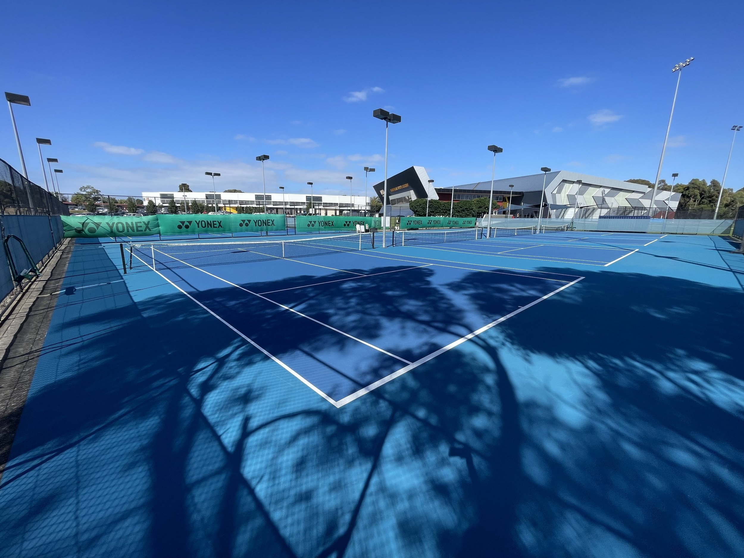 Empty blue tennis courts with white lines, surrounded by green windbreaks with "Yonex" branding, under a blue sky with some clouds and shadows from nearby trees.