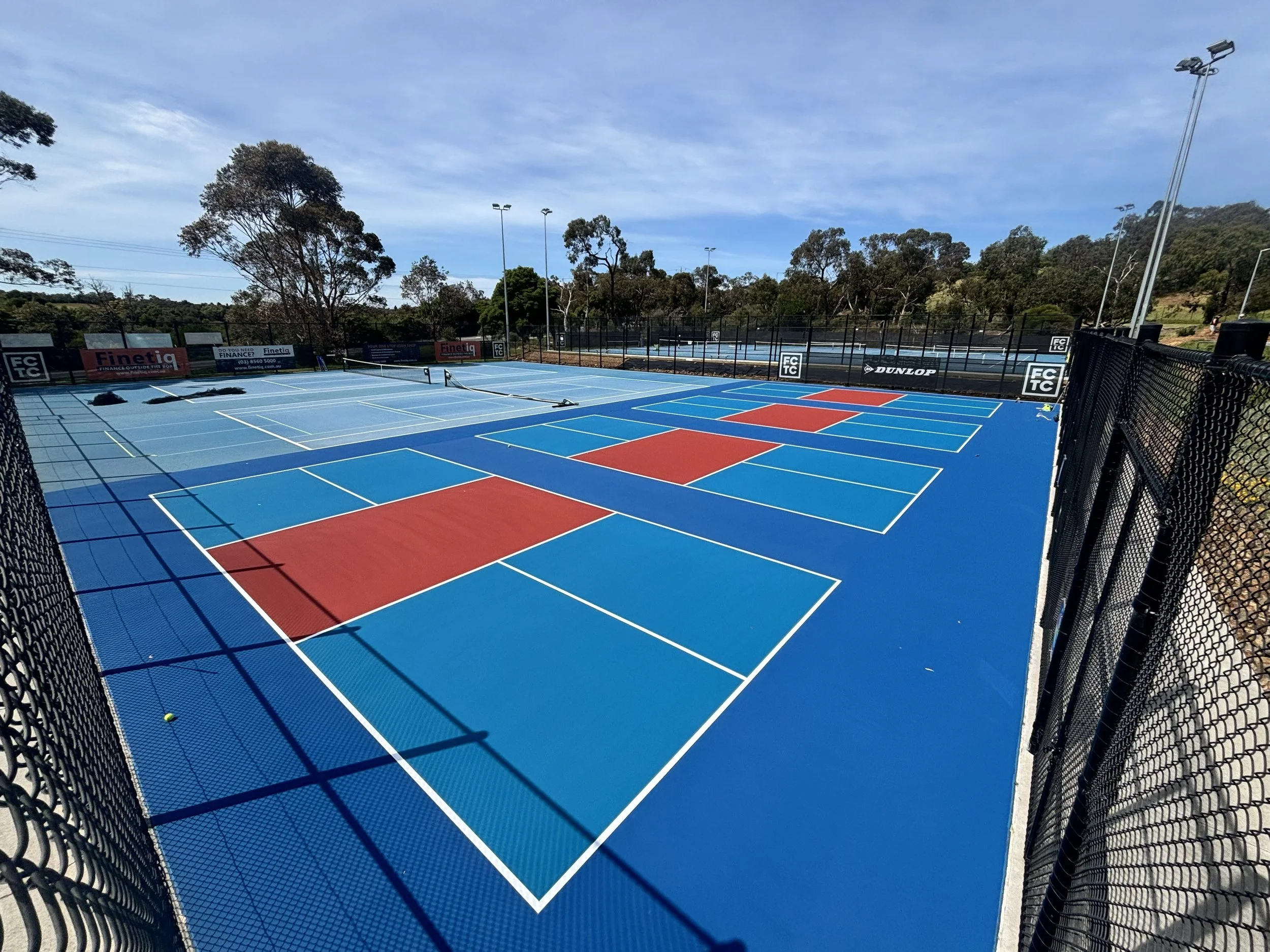 Blue tennis courts with red and white lines, surrounded by black fences, under a partly cloudy sky with trees in the background.