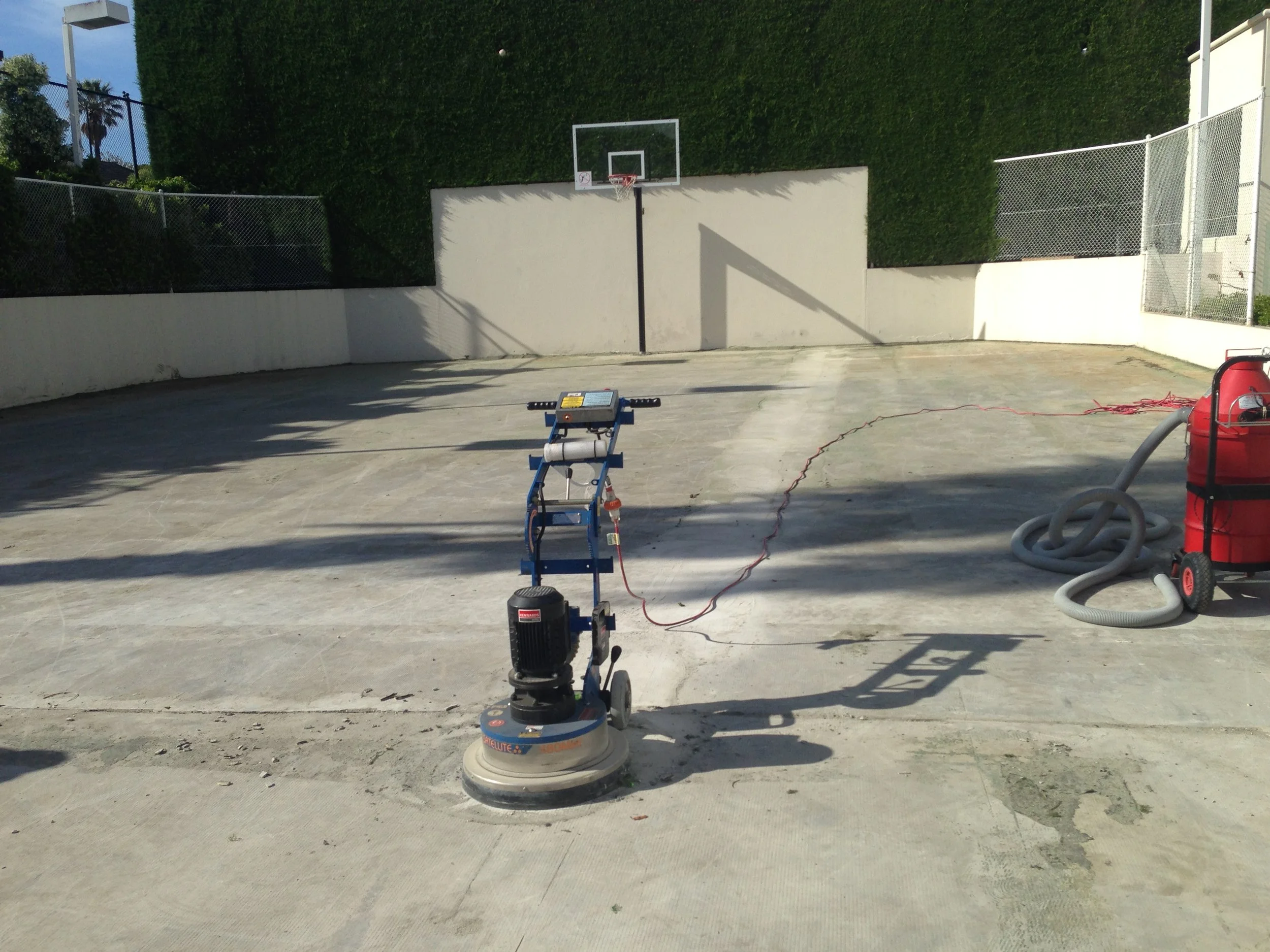 An outdoor basketball court being resurfaced with concrete. There is a power trowel machine in the foreground, and a vacuum or air compressor with hoses to the right. A basketball hoop is in the background, and the court is enclosed by white walls and a chain-link fence, with greenery on the perimeter.
