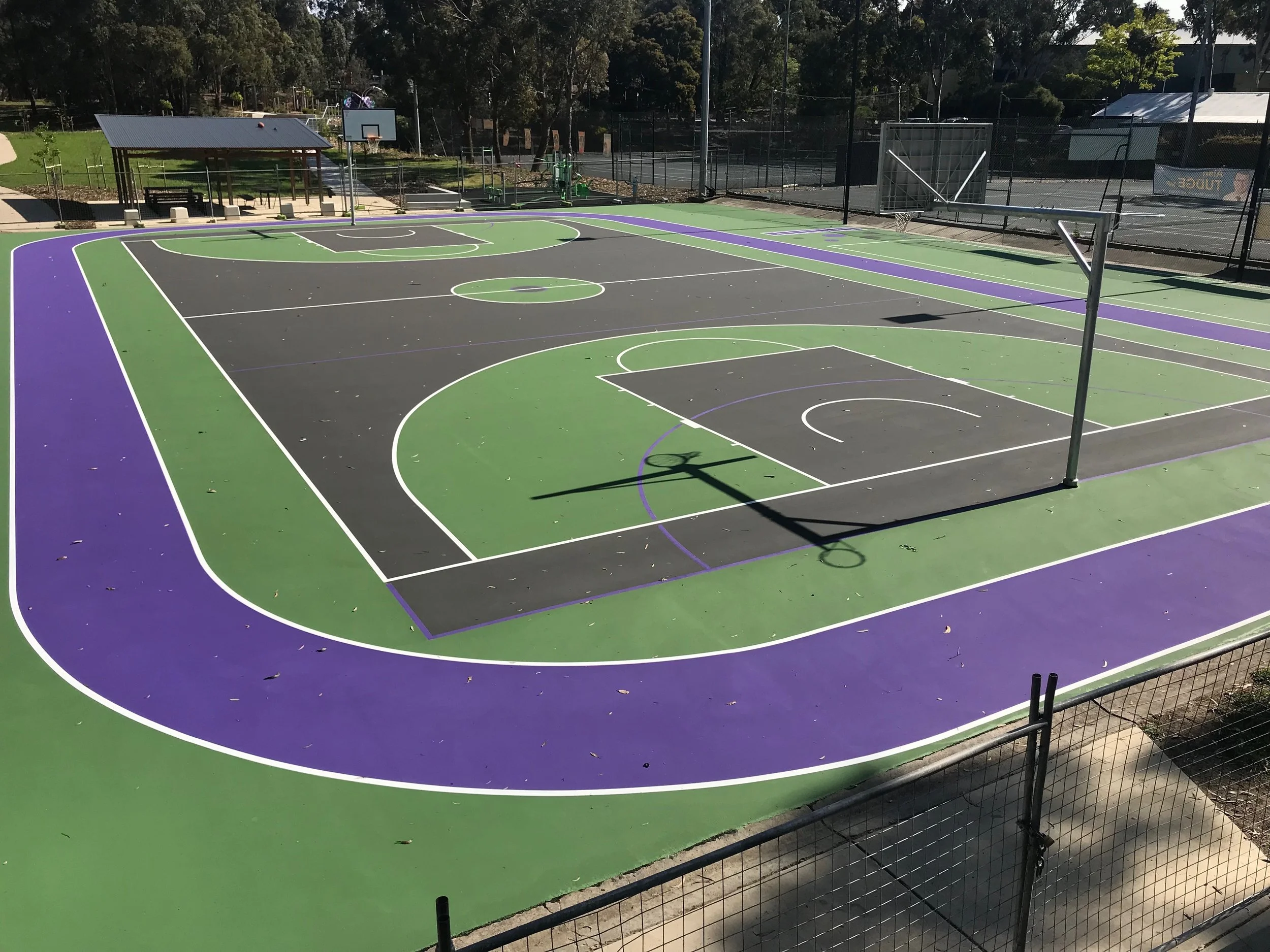 Empty outdoor basketball court with black, green, and purple surface markings, surrounded by a fence and trees in the background.