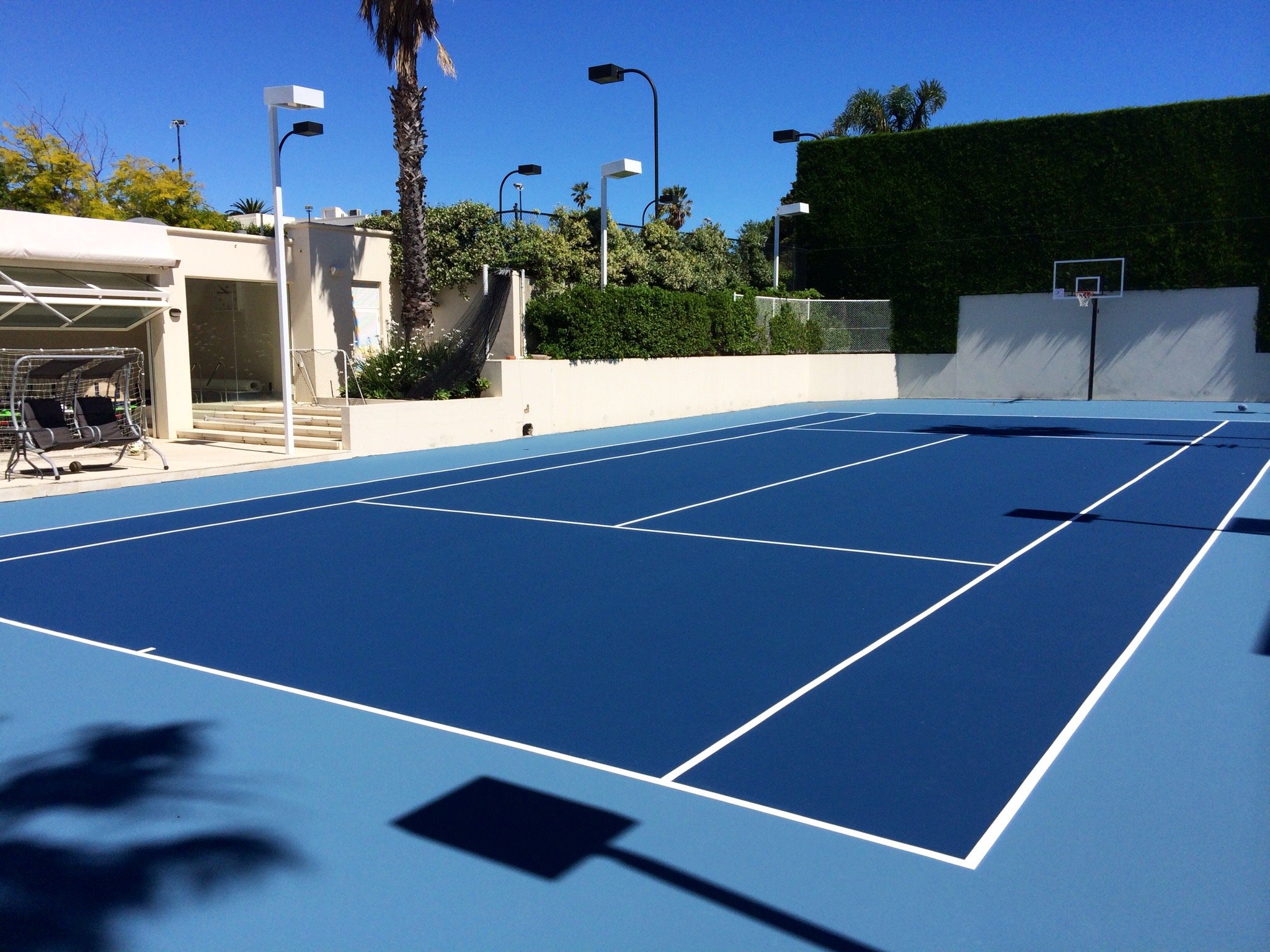 A bright blue outdoor tennis court with white lines, surrounded by trees, shrubs, and a white wall, under a clear blue sky.