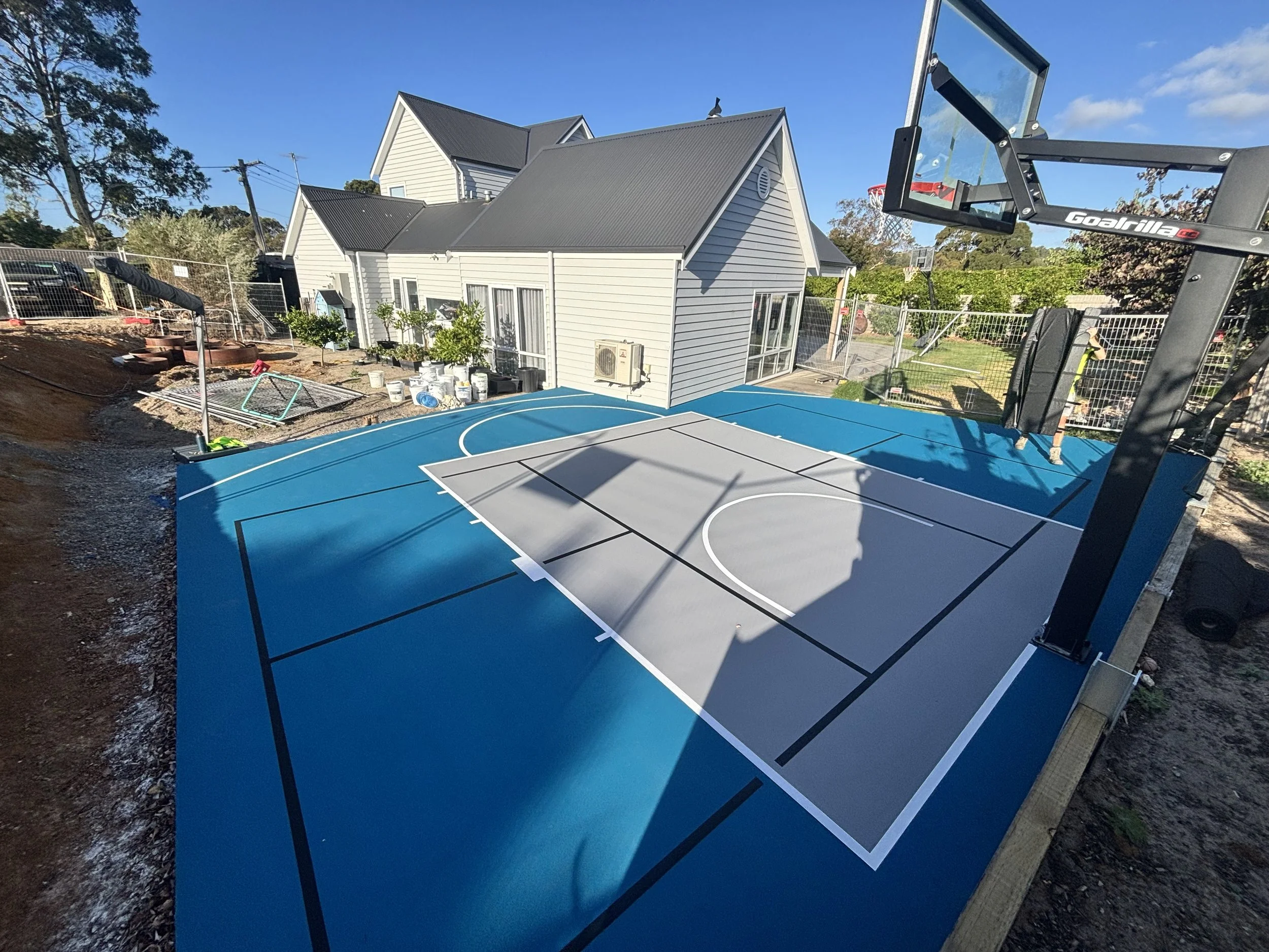Newly installed outdoor basketball court with blue and gray surface, white and black lines, and backboard, in a residential backyard with houses and greenery in the background.