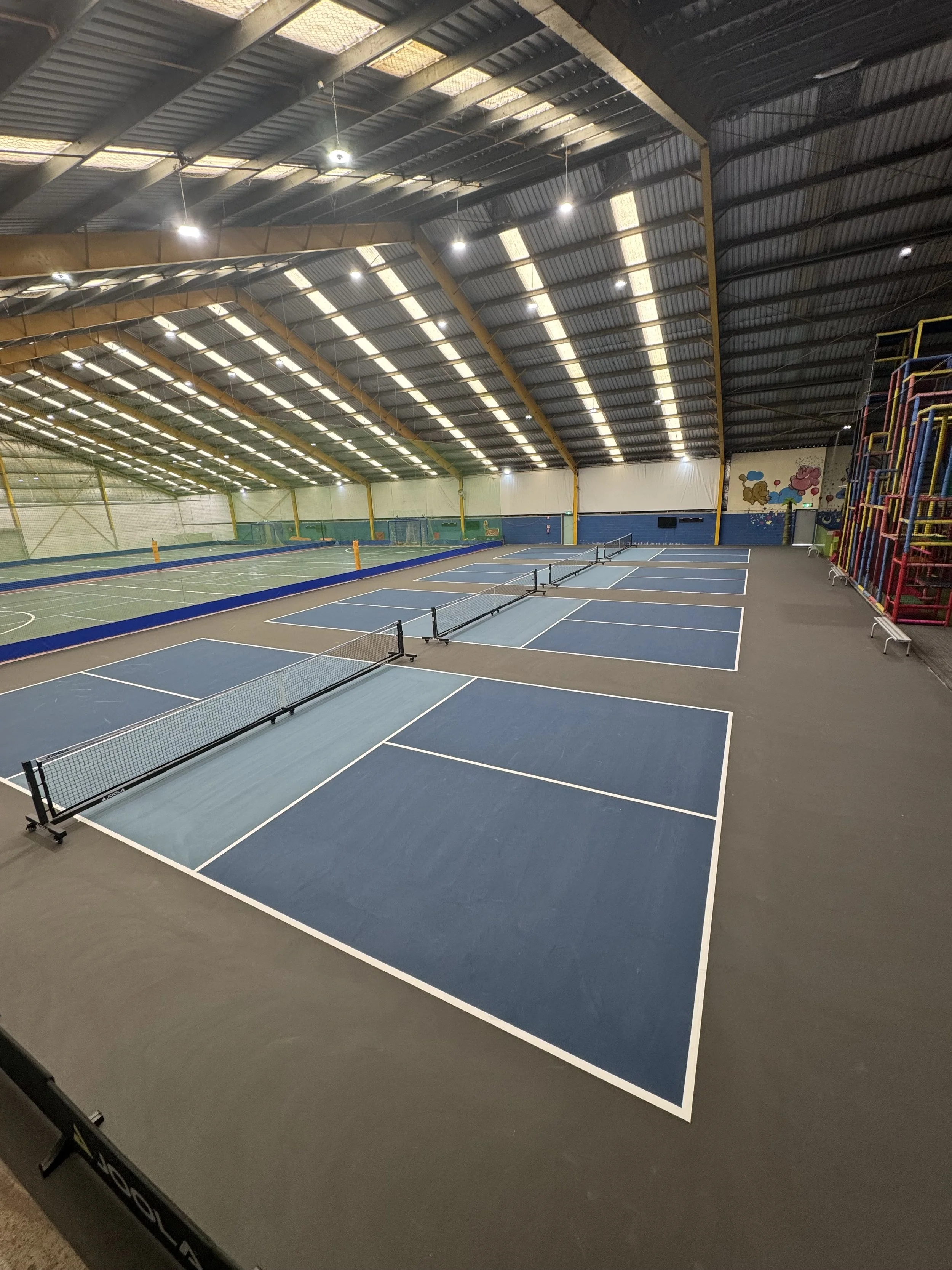 Indoor sports facility with multiple tennis and pickleball courts, playing nets, and colorful climbing equipment on the side, under a large metal roof with lighting.