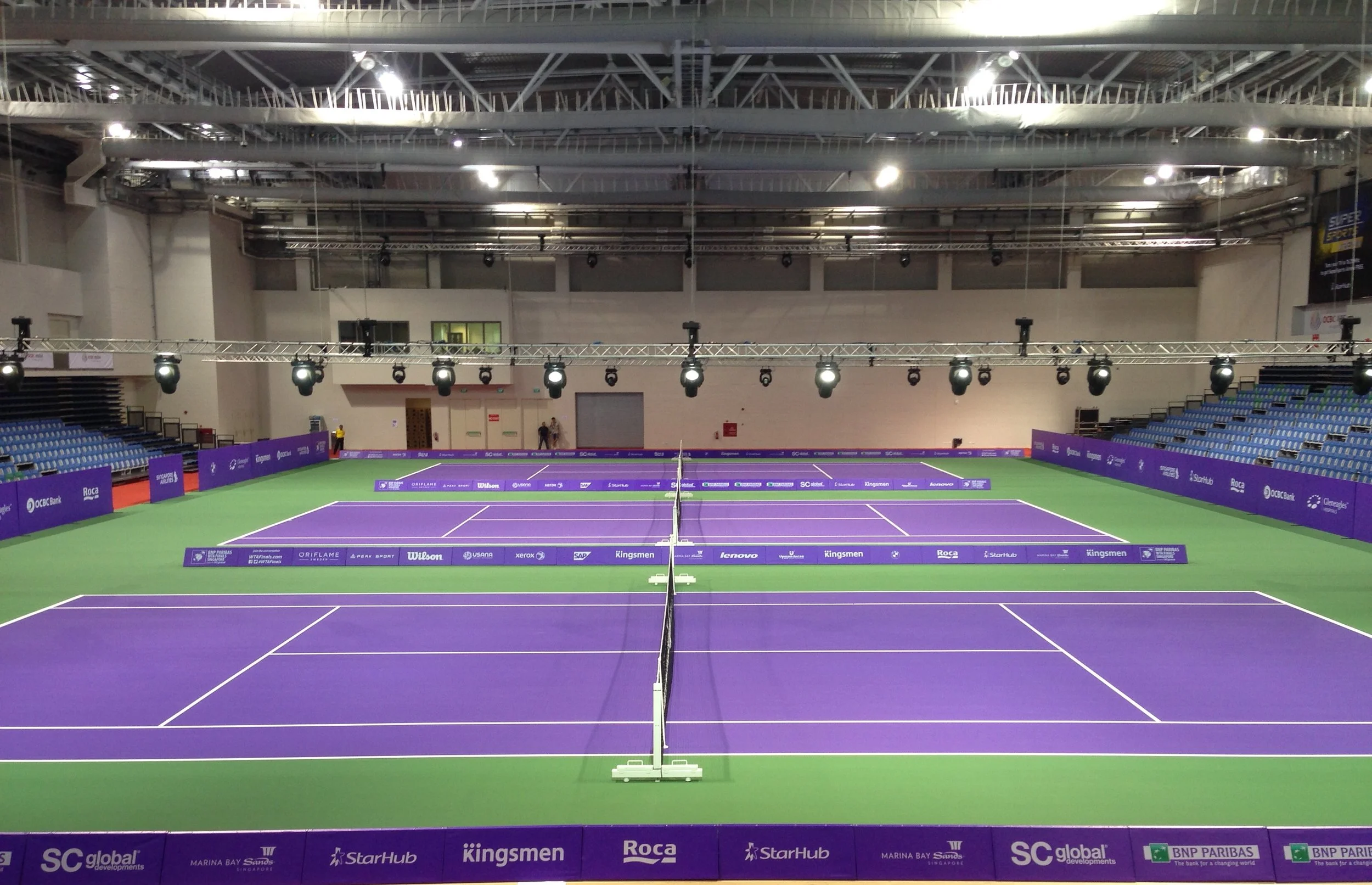 Empty indoor tennis court with purple and green surface, surrounded by blue seating and purple advertising banners, with lighting fixtures above.