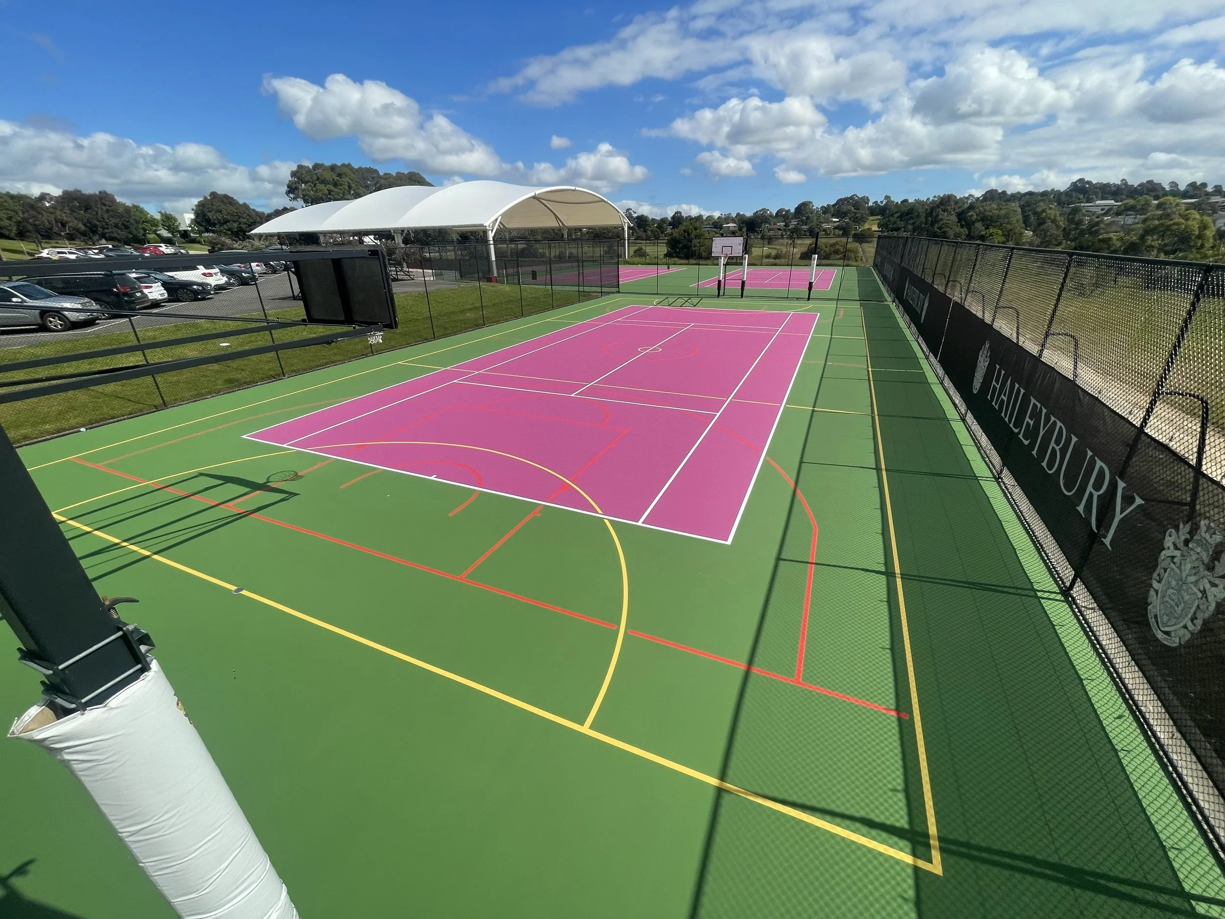 Colorful outdoor sports court with pink, green, white, yellow, and red lines, surrounded by a black fence with 'Haileybury' signage, under a partly cloudy sky.