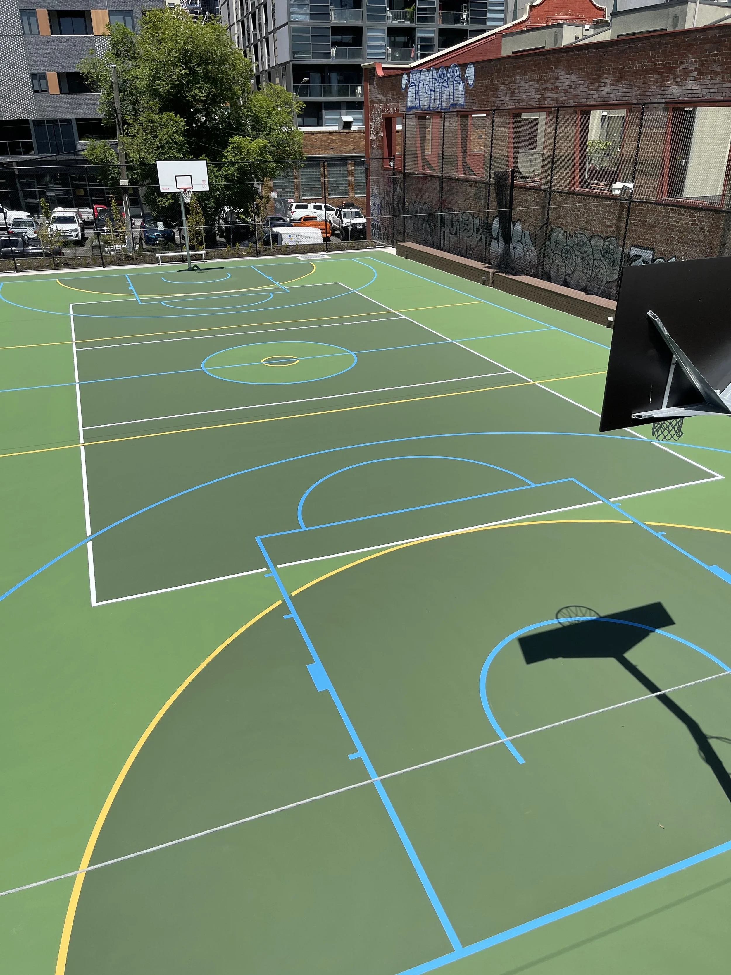 An outdoor basketball court with colorful painted lines for various sports, surrounded by a black fence, urban buildings, and parked cars.