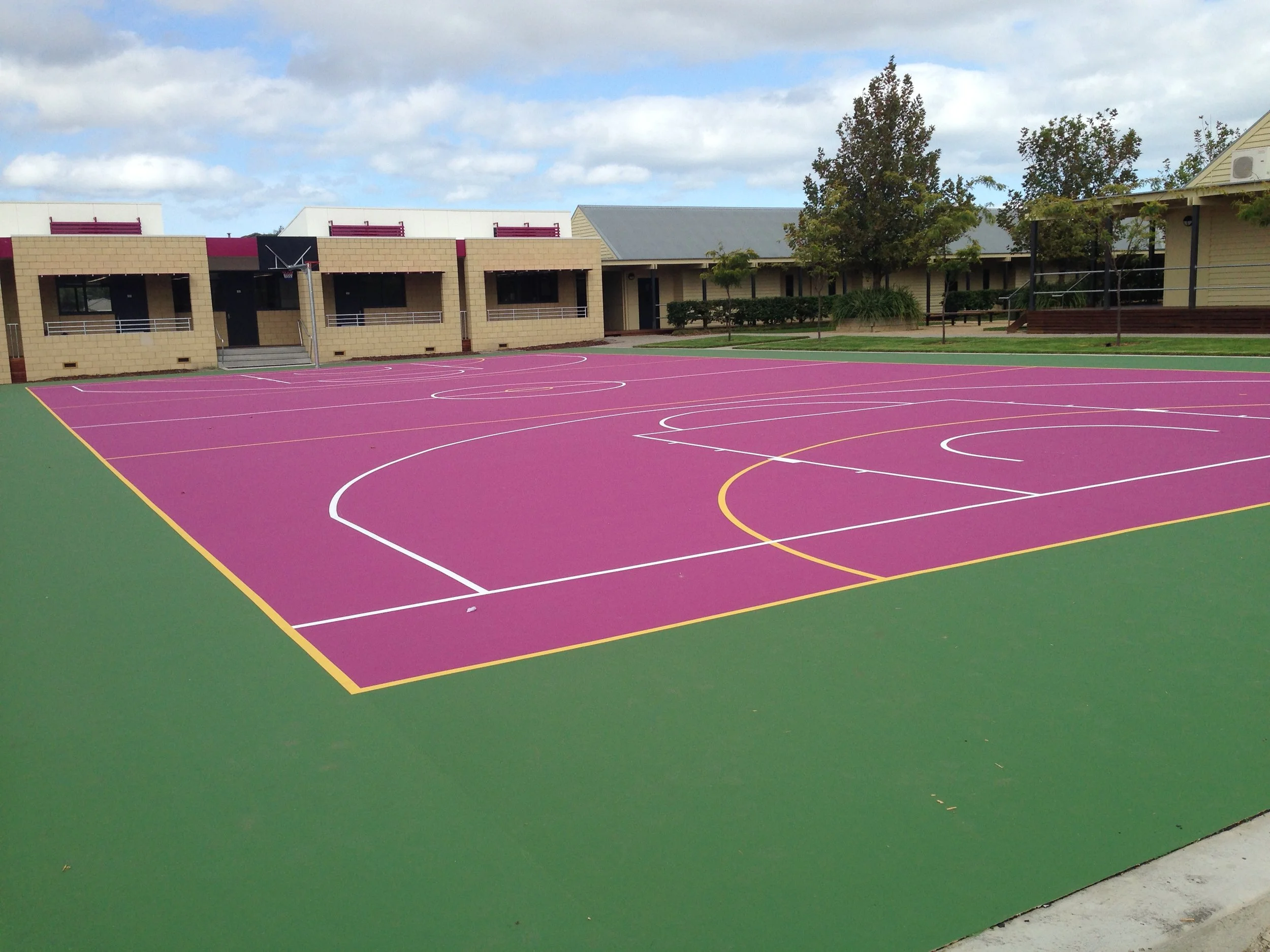 Colorful outdoor basketball court with purple playing surface and yellow, white, and green lines, surrounded by buildings, trees, and cloudy sky.