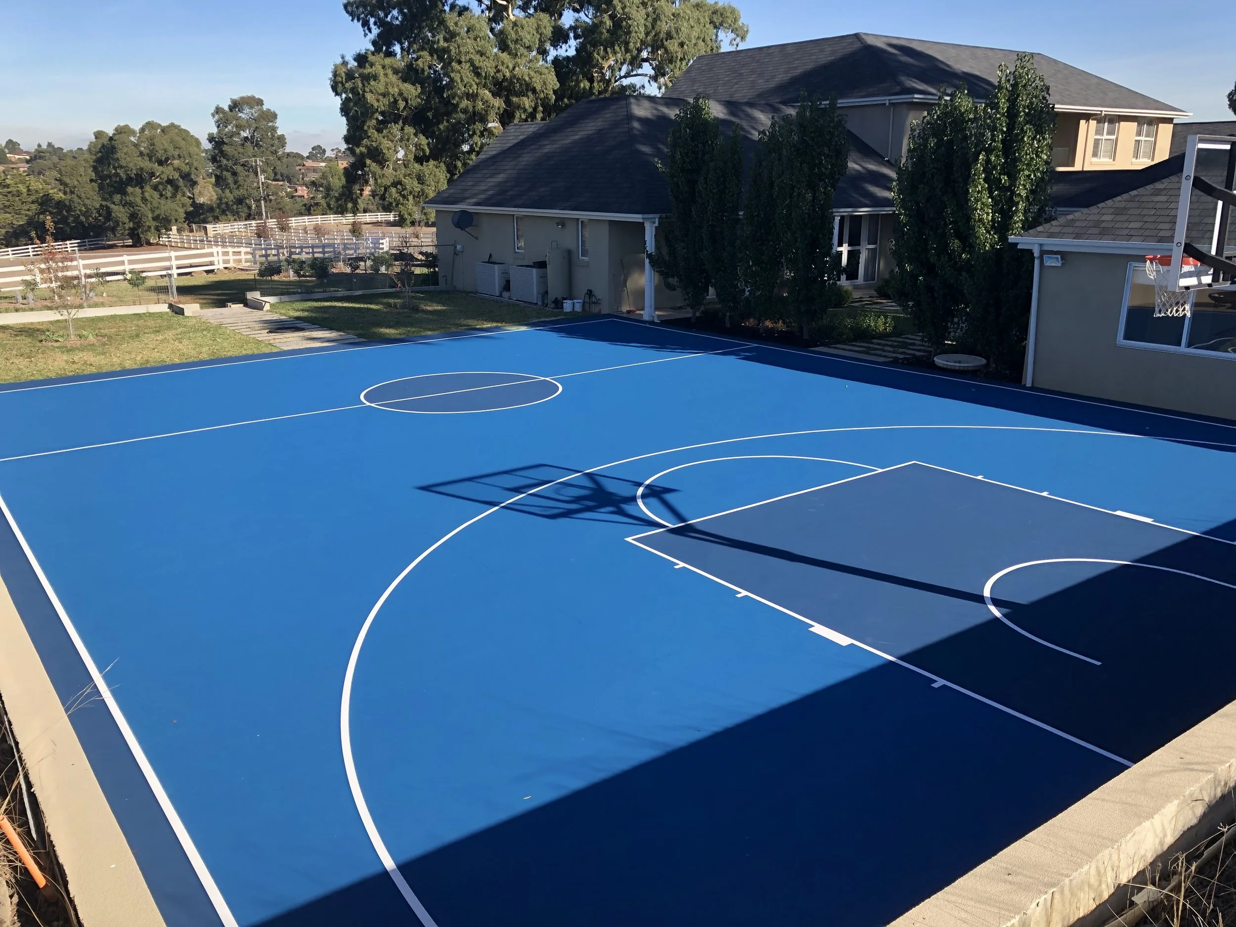 A newly painted outdoor basketball court in a residential backyard with blue surface, white court lines, basketball hoops, surrounded by trees, houses, and clear sky.