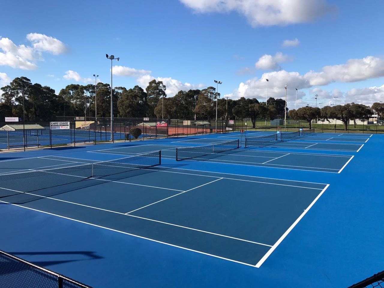 Multiple blue tennis courts outdoors on a sunny day with a few players in the distance, surrounded by a fence and trees.