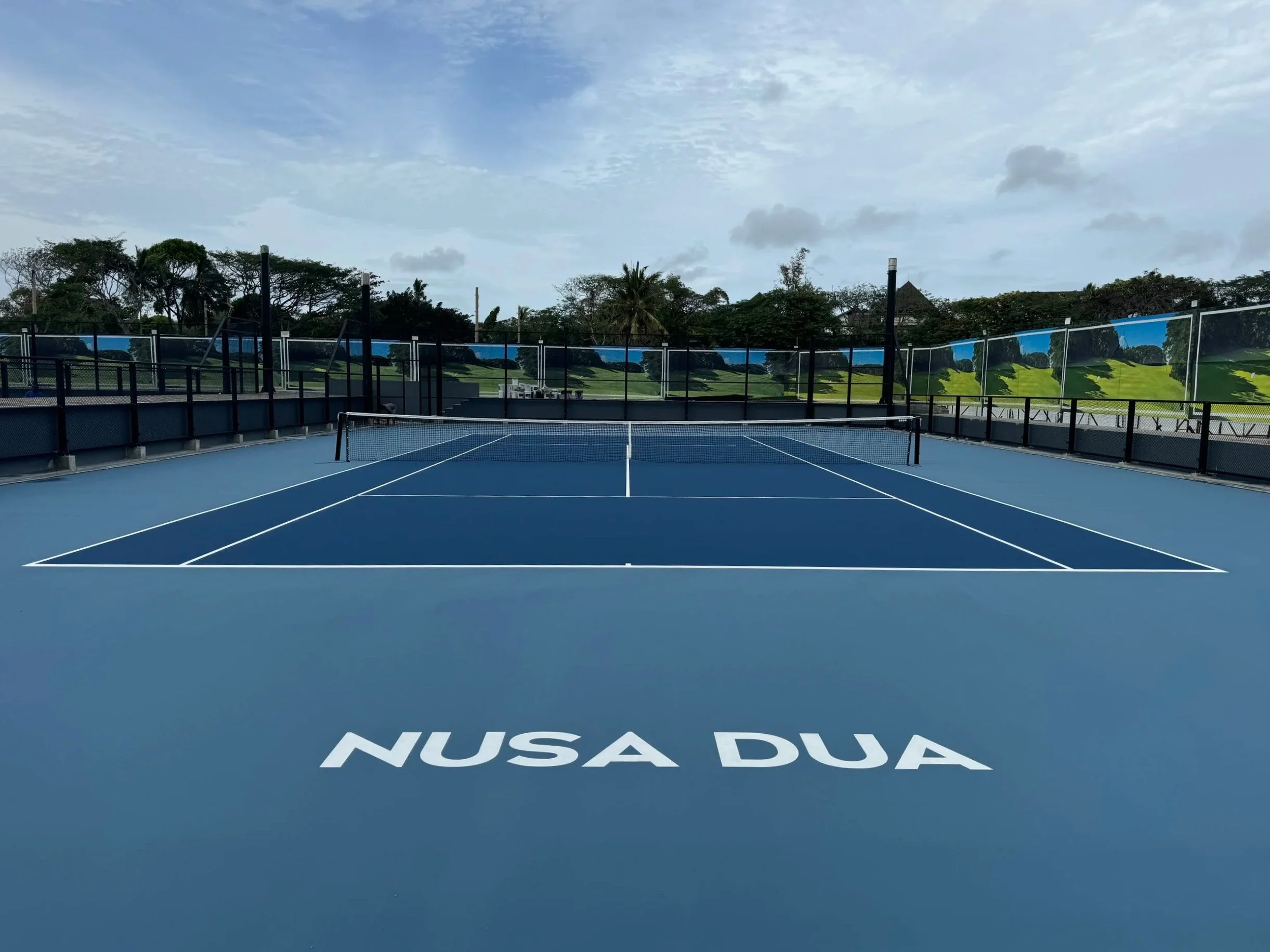 Empty blue outdoor tennis court with a net in the center, surrounded by a black fence and scenic trees and sky in the background.